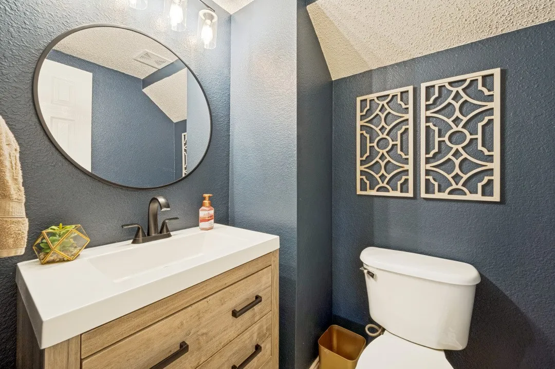 Wood-finish vanity with a white countertop sink and matte black faucet, featuring a circular mirror and overhead lighting
