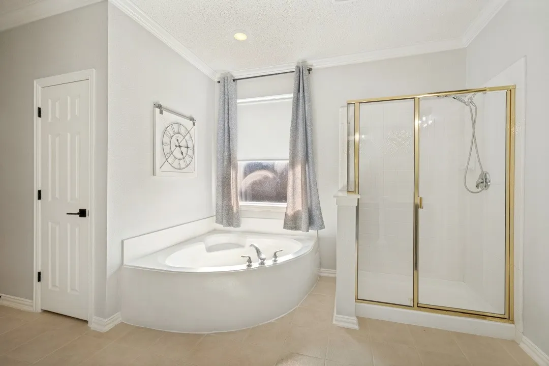 Corner soaking tub with a window, tile flooring, and an enclosed shower with a gold-finish frame