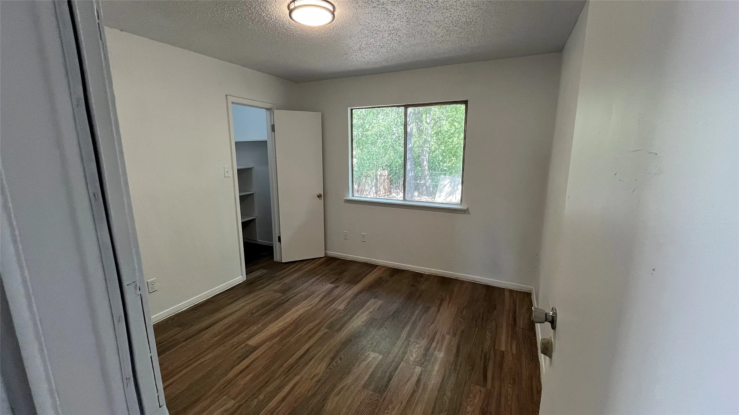 Bedroom 2 - Wood-finish flooring, overhead flush mount light fixture, window with exterior foliage, and built-in closet shelving