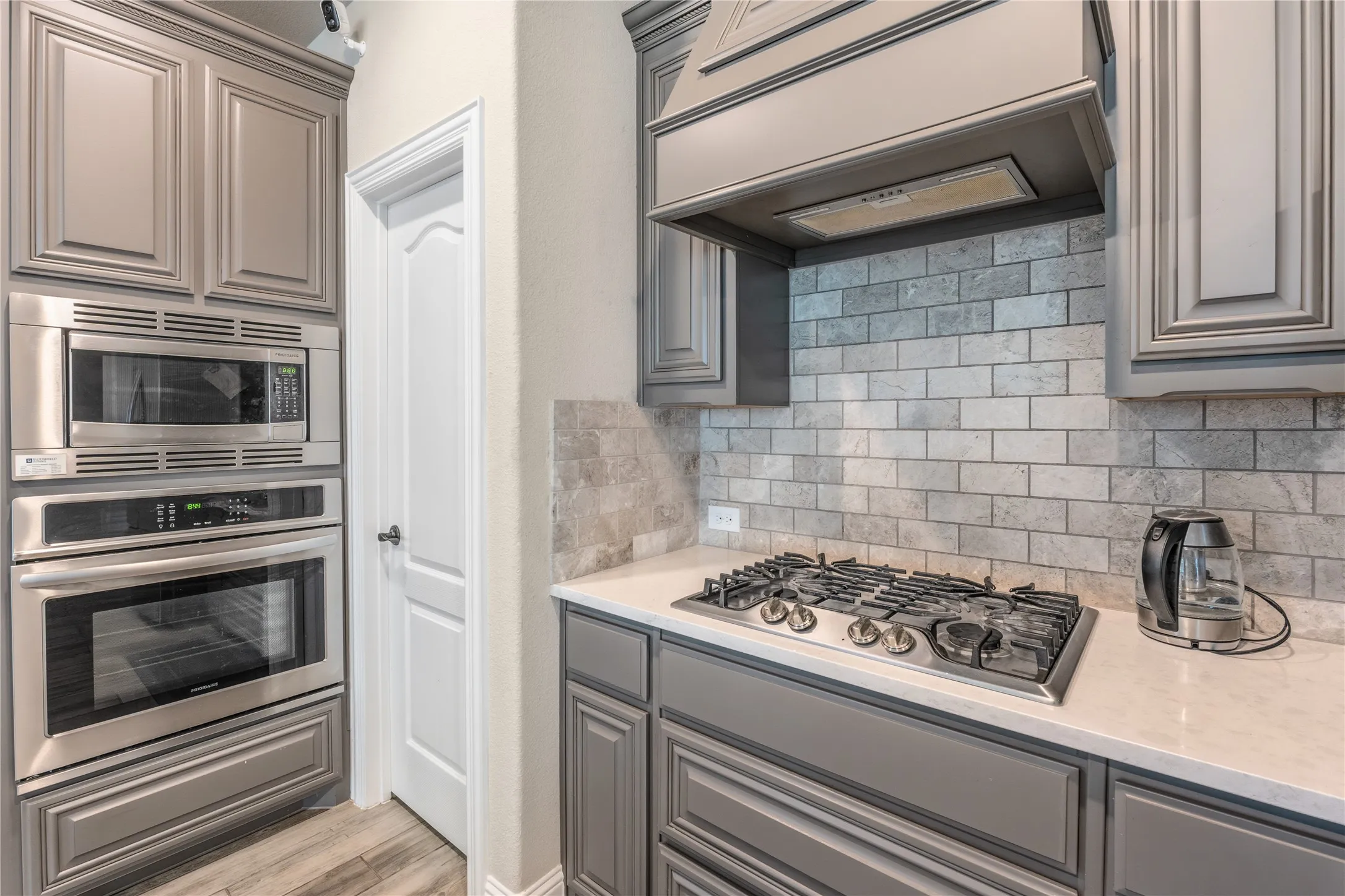 Kitchen with gray cabinets, stainless steel appliances, light stone countertops, backsplash, and light wood-style flooring