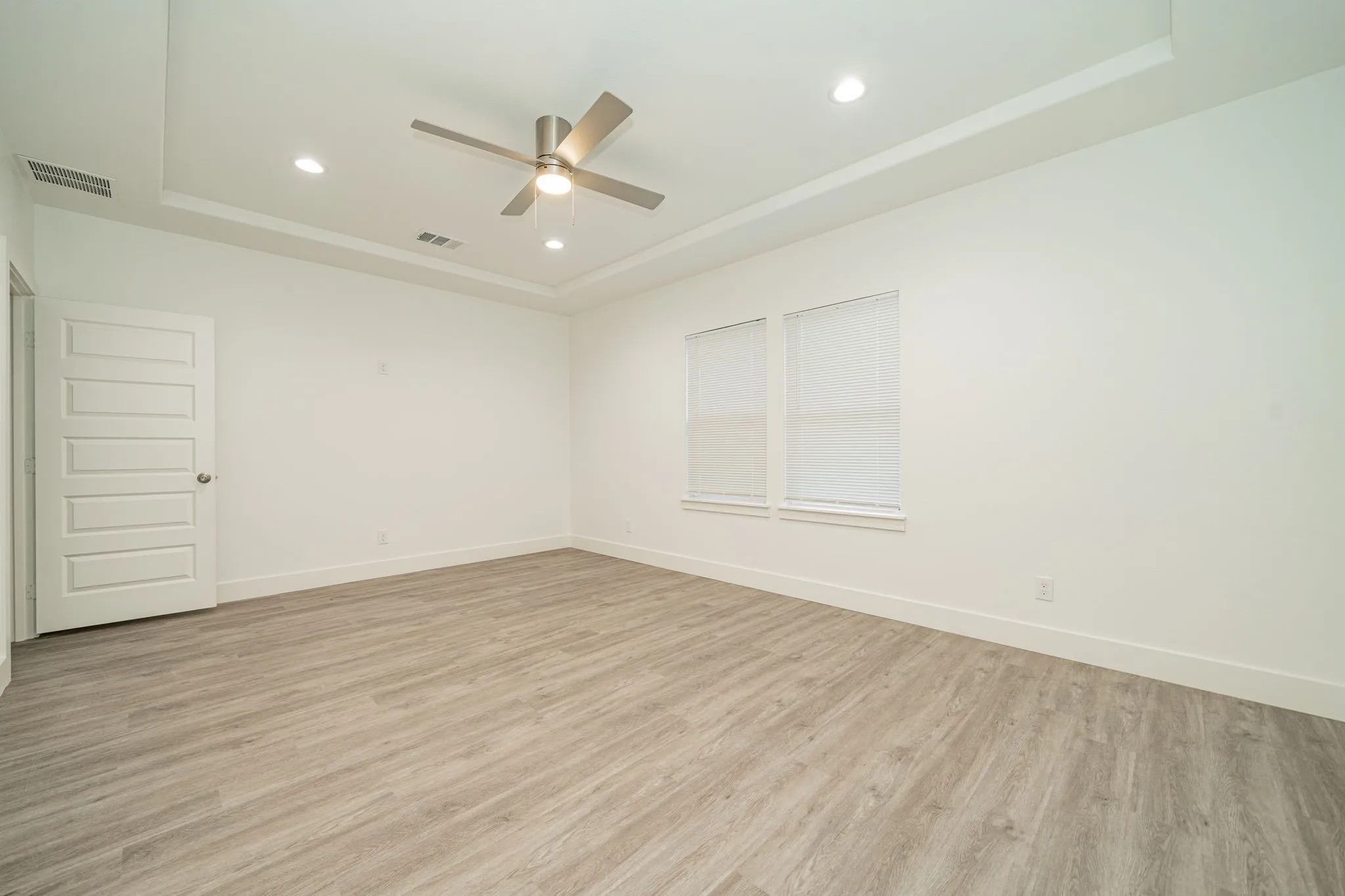 Tray ceiling with recessed lighting, modern ceiling fan, wood-finish flooring, two windows with blinds, and a paneled interior door