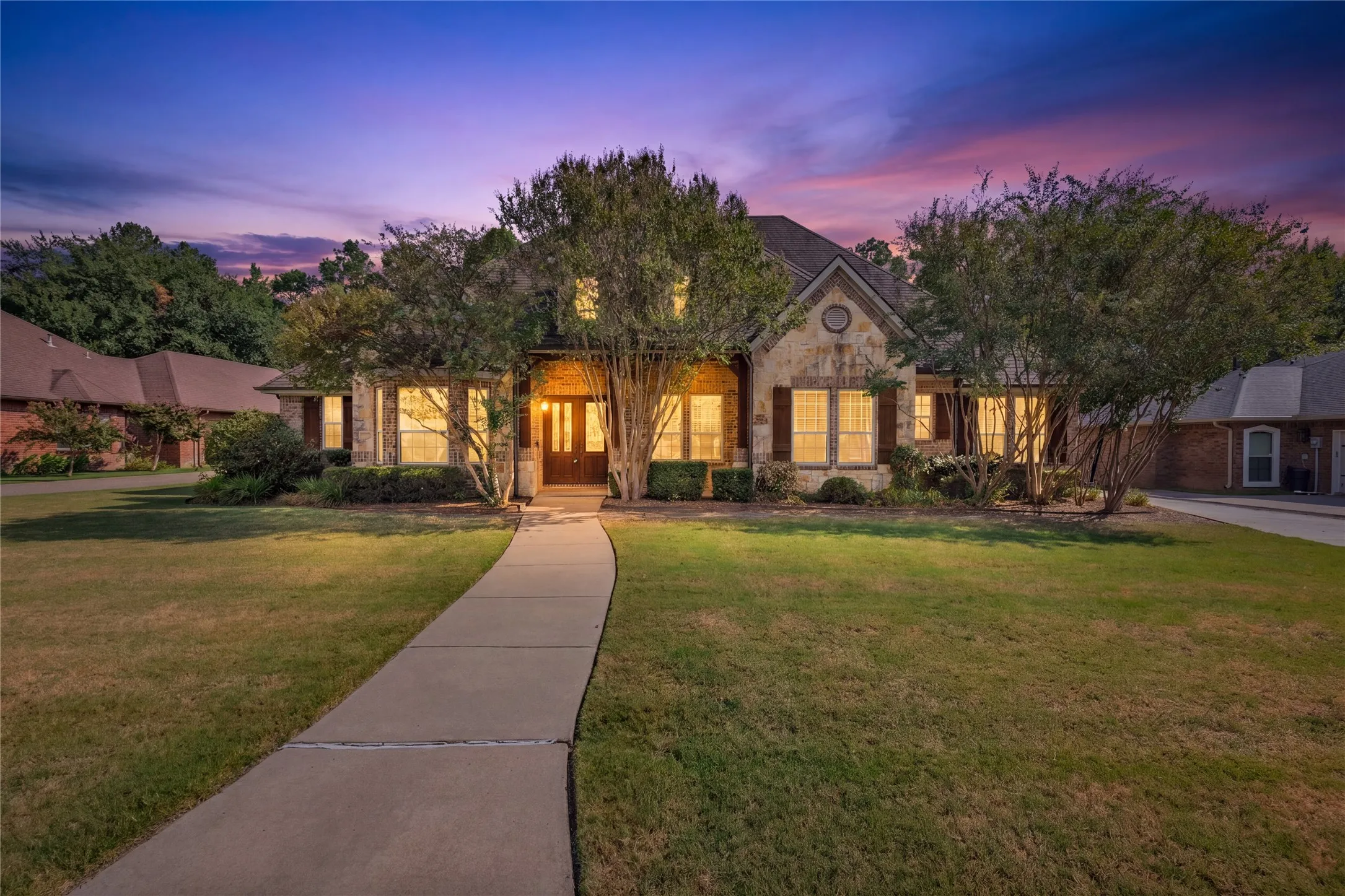 Stone facade residence featuring a front entry with double doors, multiple windows with shutters, a curved concrete pathway, and mature landscaping