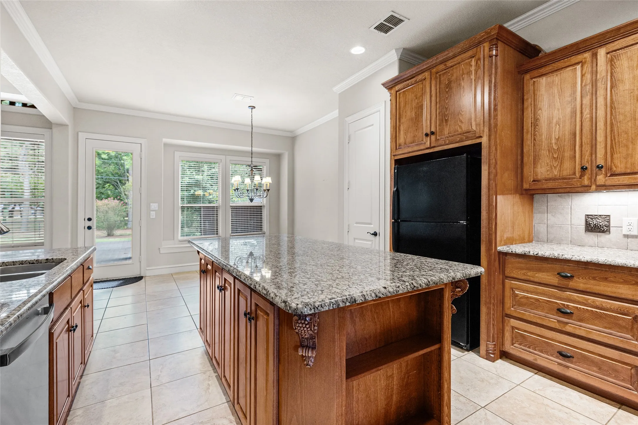 Spacious kitchen featuring warm wood cabinetry, granite countertops, a large center island with seating, stainless steel appliances, tile flooring, recessed lighting, and abundant natural light from multiple windows and glass doors overlooking the outdoor space