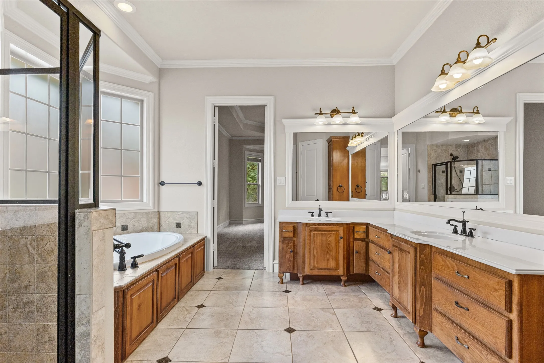 Double vanity with wood-finish cabinetry, white countertops, dual mirrors with ornate lighting, and a built-in soaking tub with tile surround