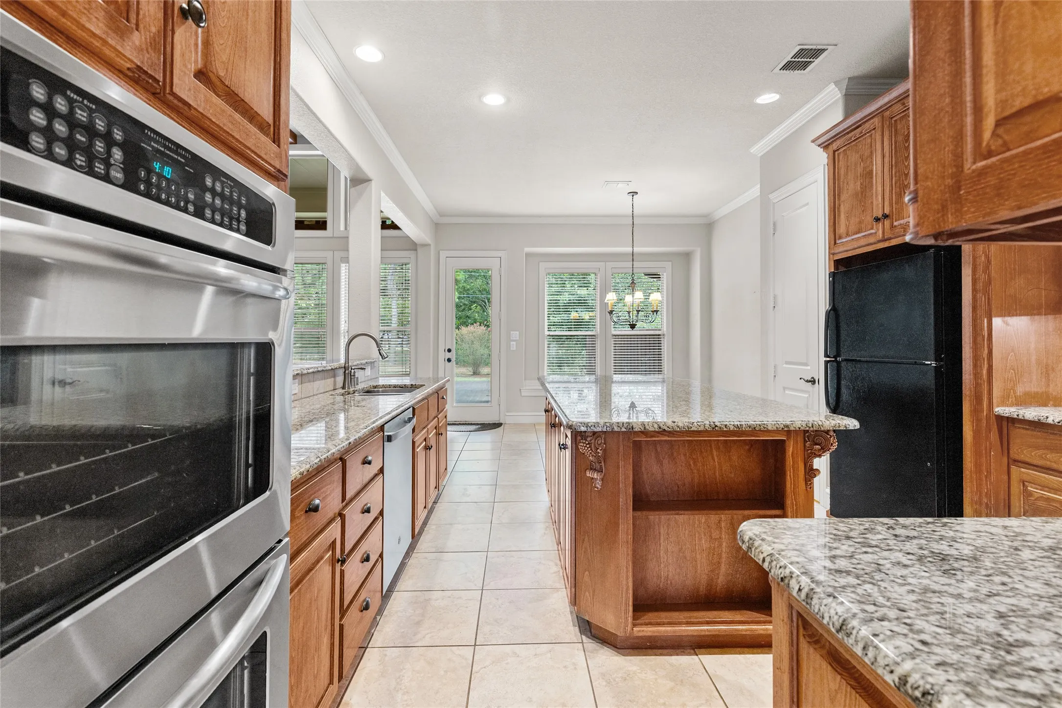 Galley kitchen featuring stainless steel dual wall ovens, wood cabinetry with multiple storage drawers, granite countertops, tile flooring, and abundant natural light from multi-pane windows and glass doors opening to outdoor space