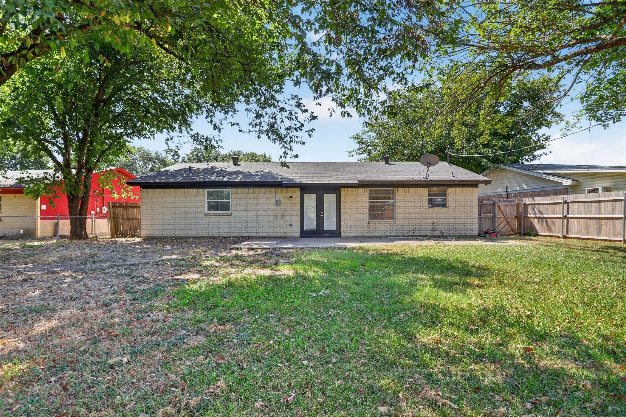 Expansive backyard featuring a brick exterior, a concrete patio, wood fencing, and mature trees providing natural shade
