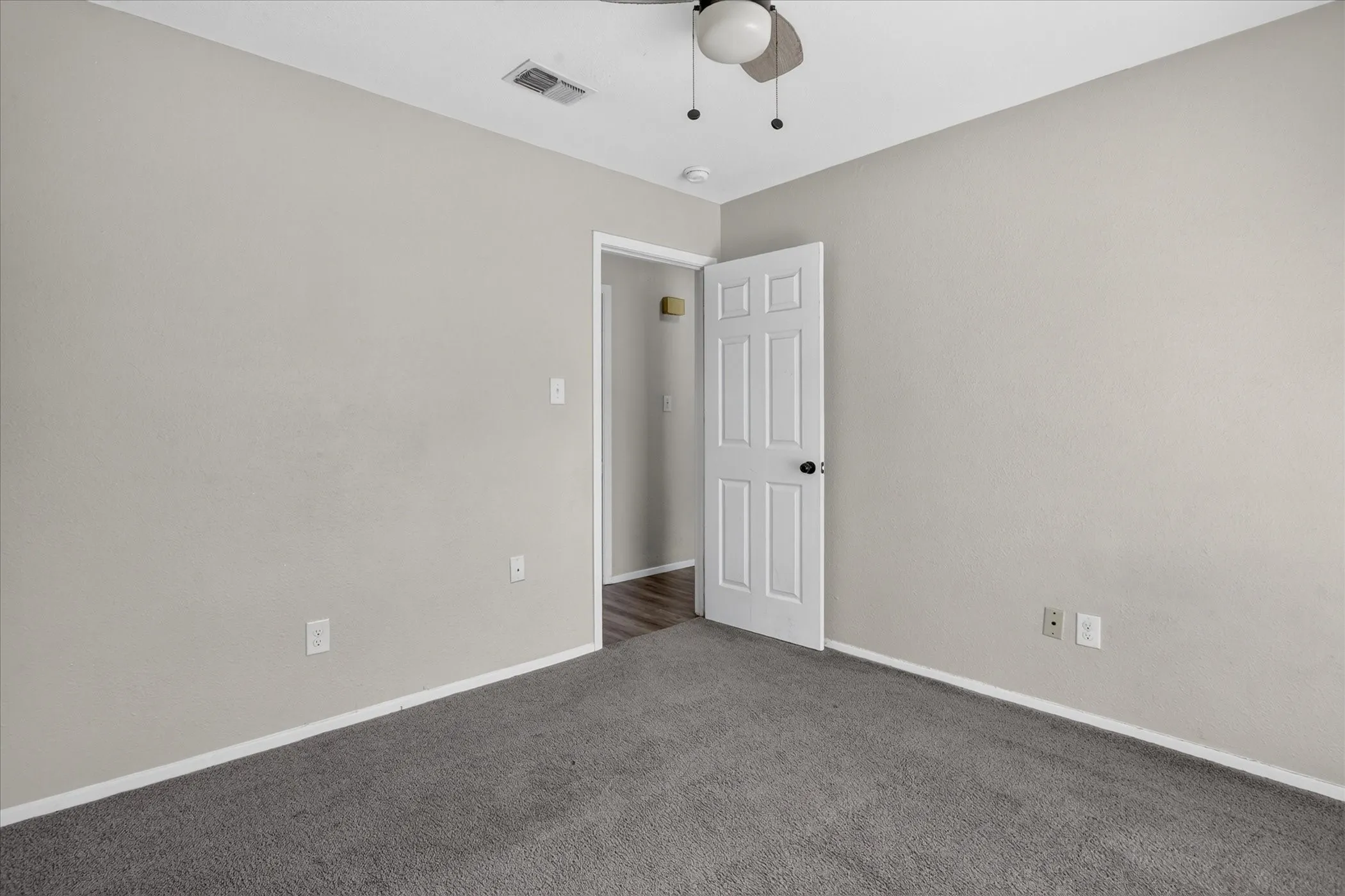 Neutral-toned walls, grey carpeting, ceiling fan, white paneled door, and wood-finish flooring visible through the doorway