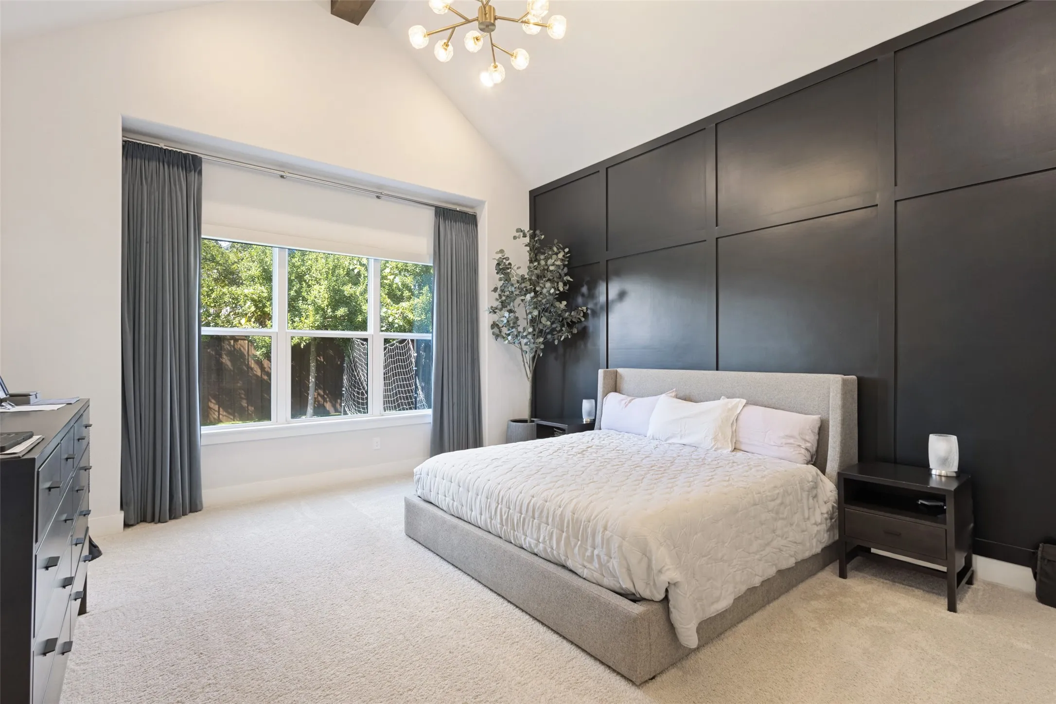 Vaulted ceiling with an attached light fixture, a large window with privacy shades, a dark paneled accent wall, and light-colored carpeting