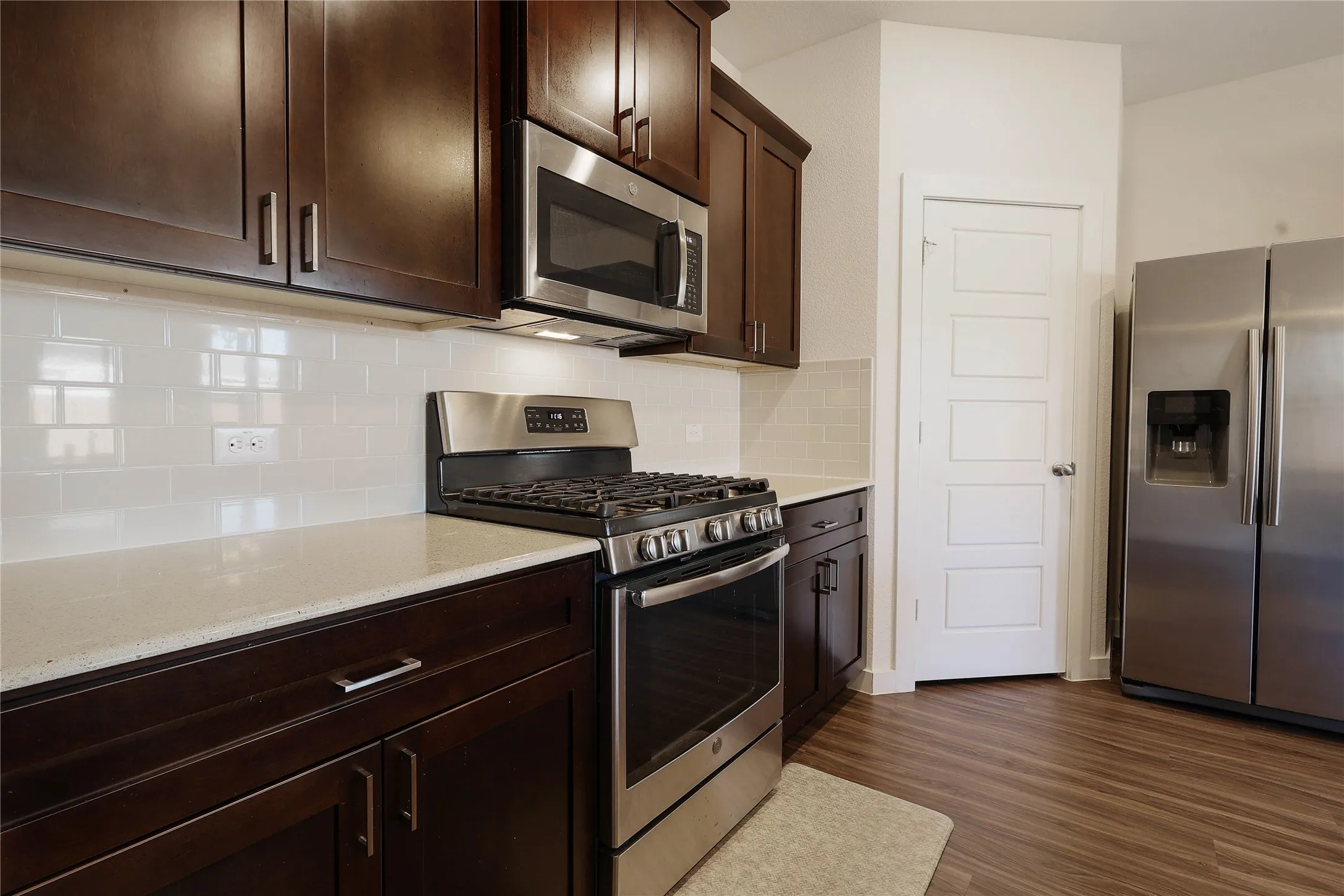 Stainless steel appliances, rich wood-finish cabinetry, light-toned countertops, white subway tile backsplash, and wood-finish flooring