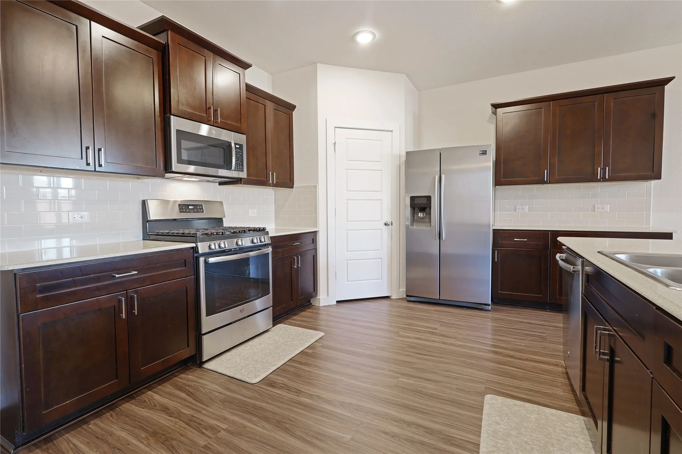 Contemporary kitchen featuring wood-finish flooring, dark wood cabinetry, stainless steel appliances, a white subway tile backsplash, and light-toned countertops