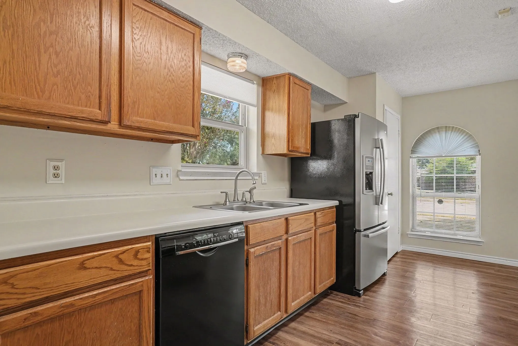 Wood-finish flooring, wood cabinetry, white countertops, dual basin sink, and an arched window