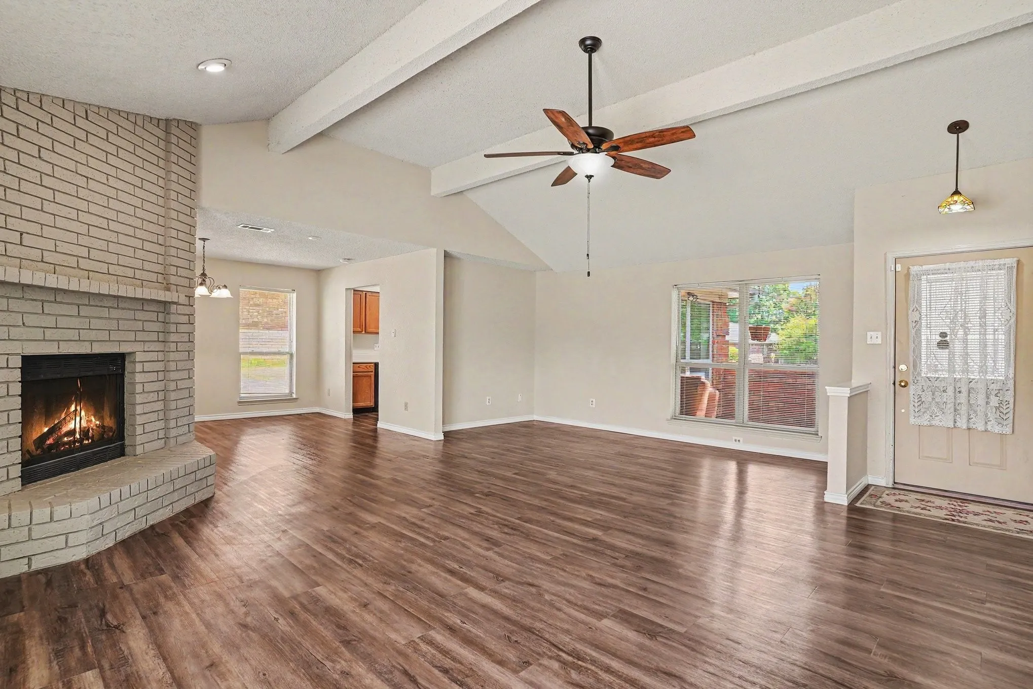 Vaulted ceiling with exposed beams, brick fireplace, wood-finish flooring, ceiling fan, and large windows