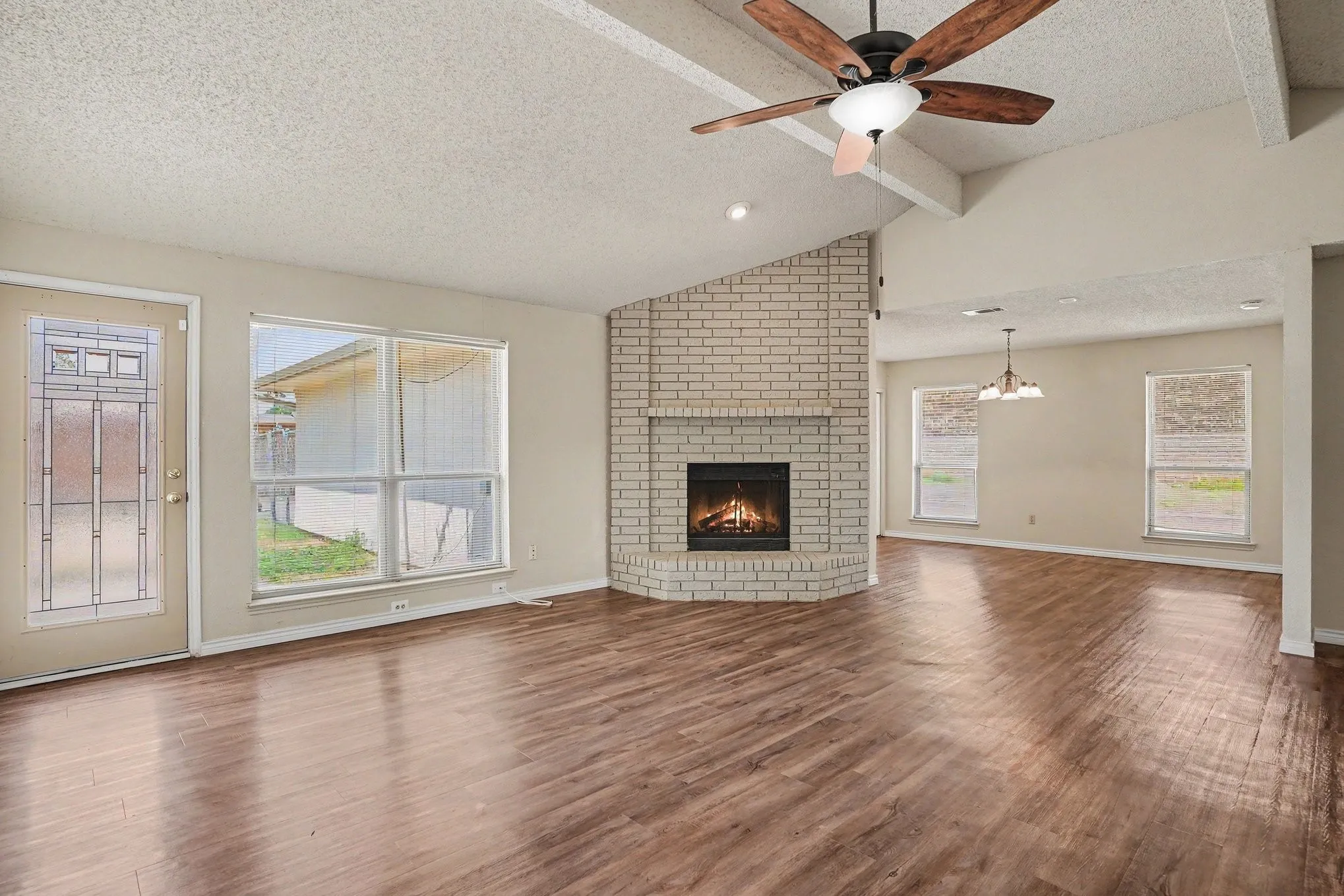 Vaulted ceiling with exposed beams, wood-finish flooring, brick fireplace, ceiling fan, and multiple windows