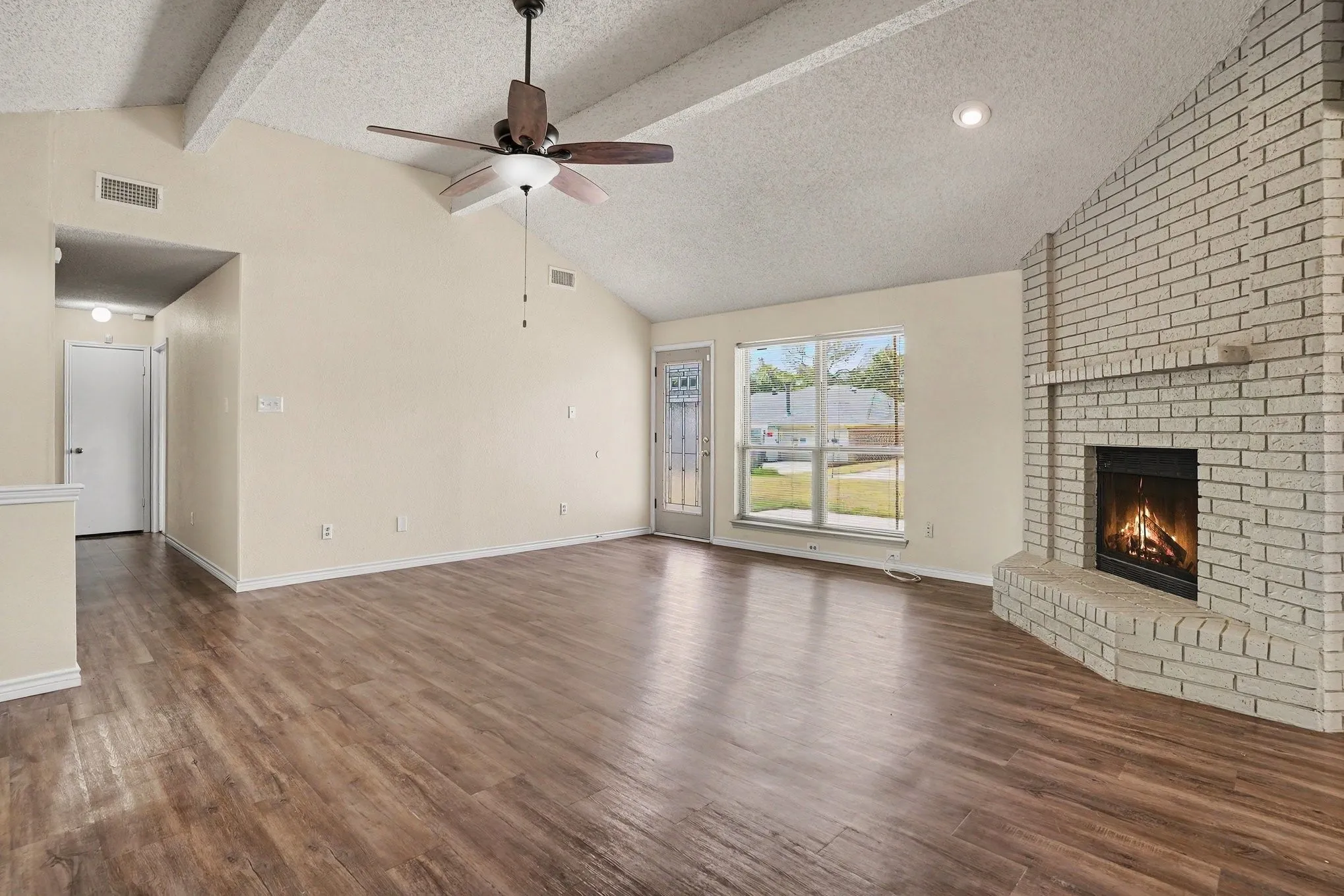 Vaulted ceiling with exposed beams, wood-finish flooring, brick fireplace, ceiling fan, and large window