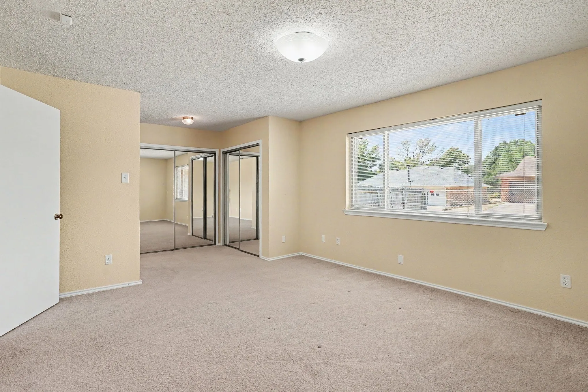 Spacious room featuring neutral carpeting, a wide window with blinds, and mirrored sliding closet doors