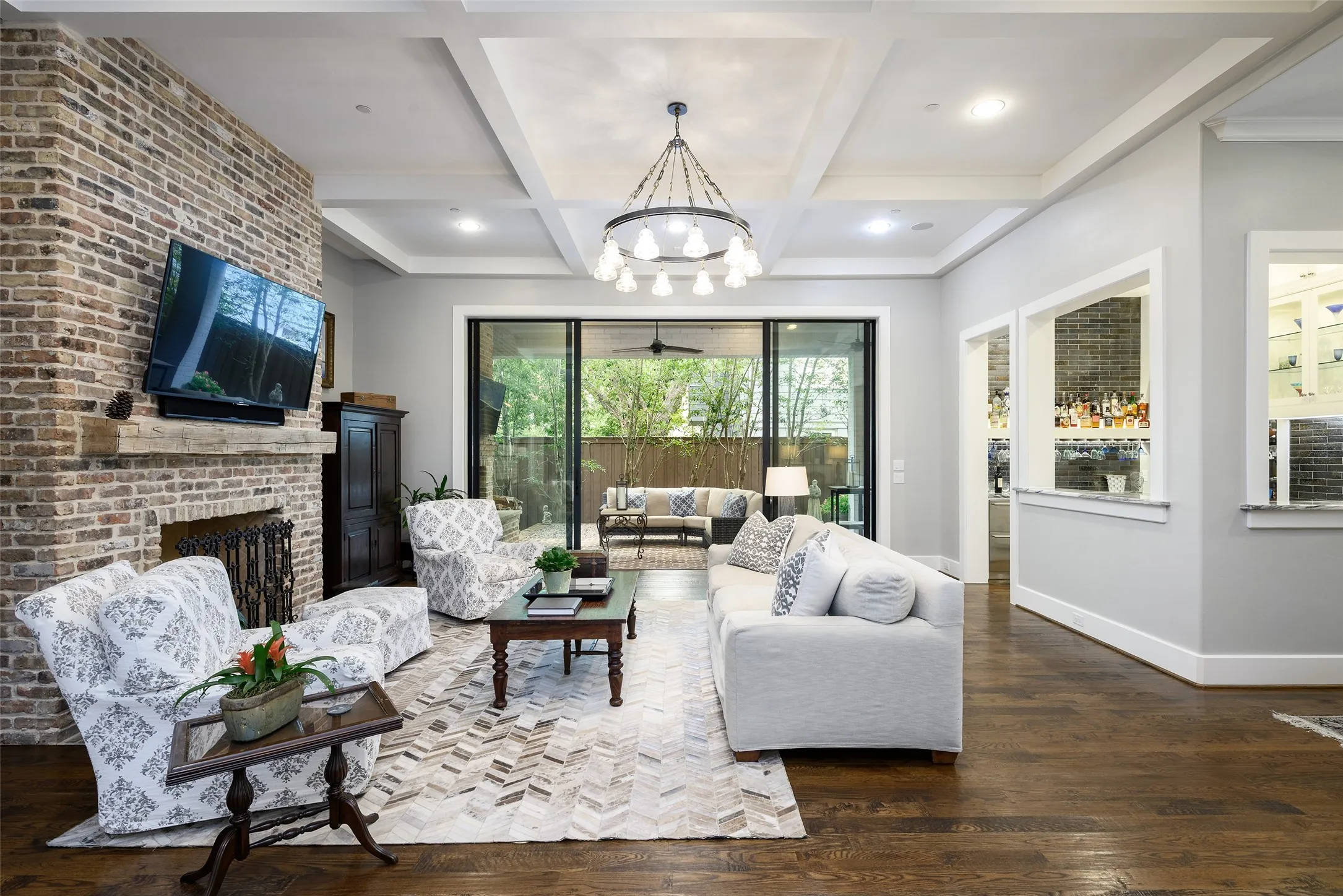 Exposed brick fireplace with a wood-beam mantel, dark wood-finish flooring, coffered ceiling, and sliding glass doors to outdoor hardscaping