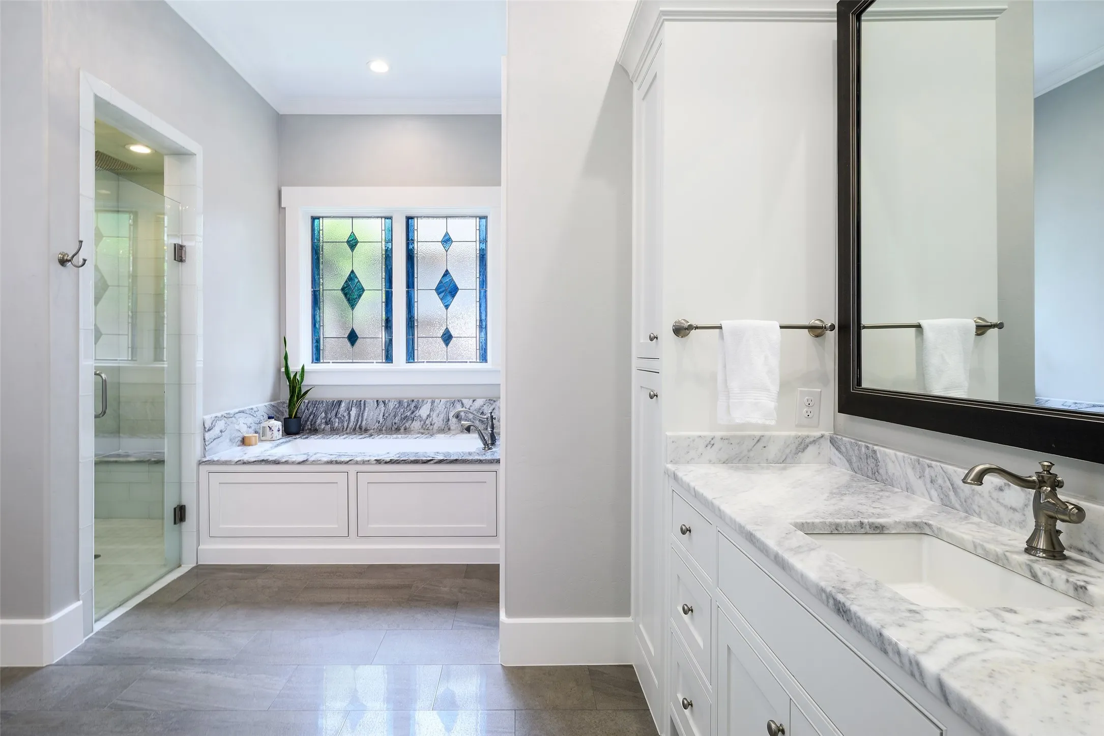 White vanity with stone countertop and integrated sink, glass-enclosed shower with tile surround, built-in storage cabinetry, and decorative stained-glass window