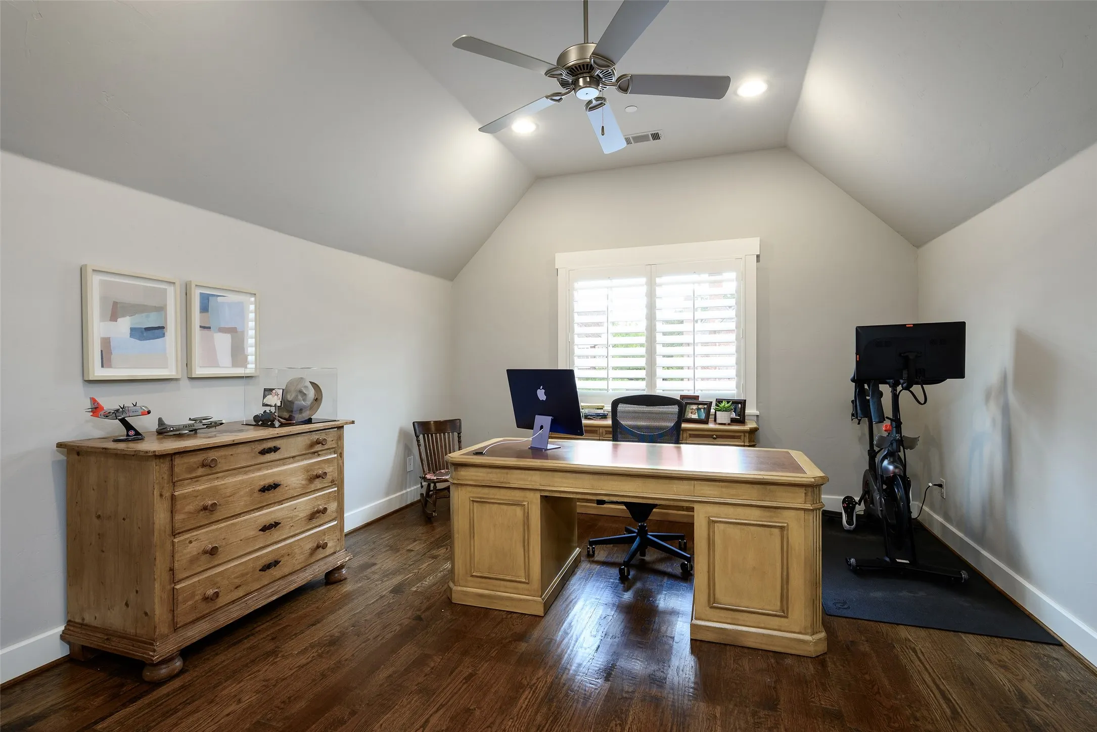 Spacious home office featuring vaulted ceilings with ceiling fan and recessed lighting, hardwood flooring, dual wooden desks with ample storage cabinetry, and shuttered windows flooding the room with natural light