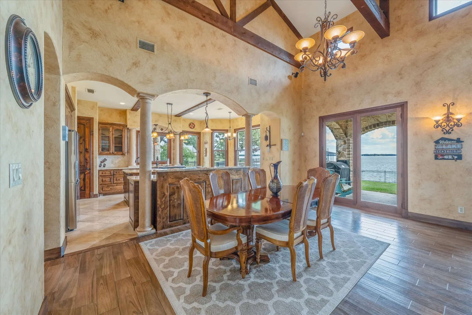 Vaulted ceiling with exposed wood beams, French doors opening to waterfront, arched doorways with decorative columns, wood-finish flooring, and built-in kitchen cabinetry