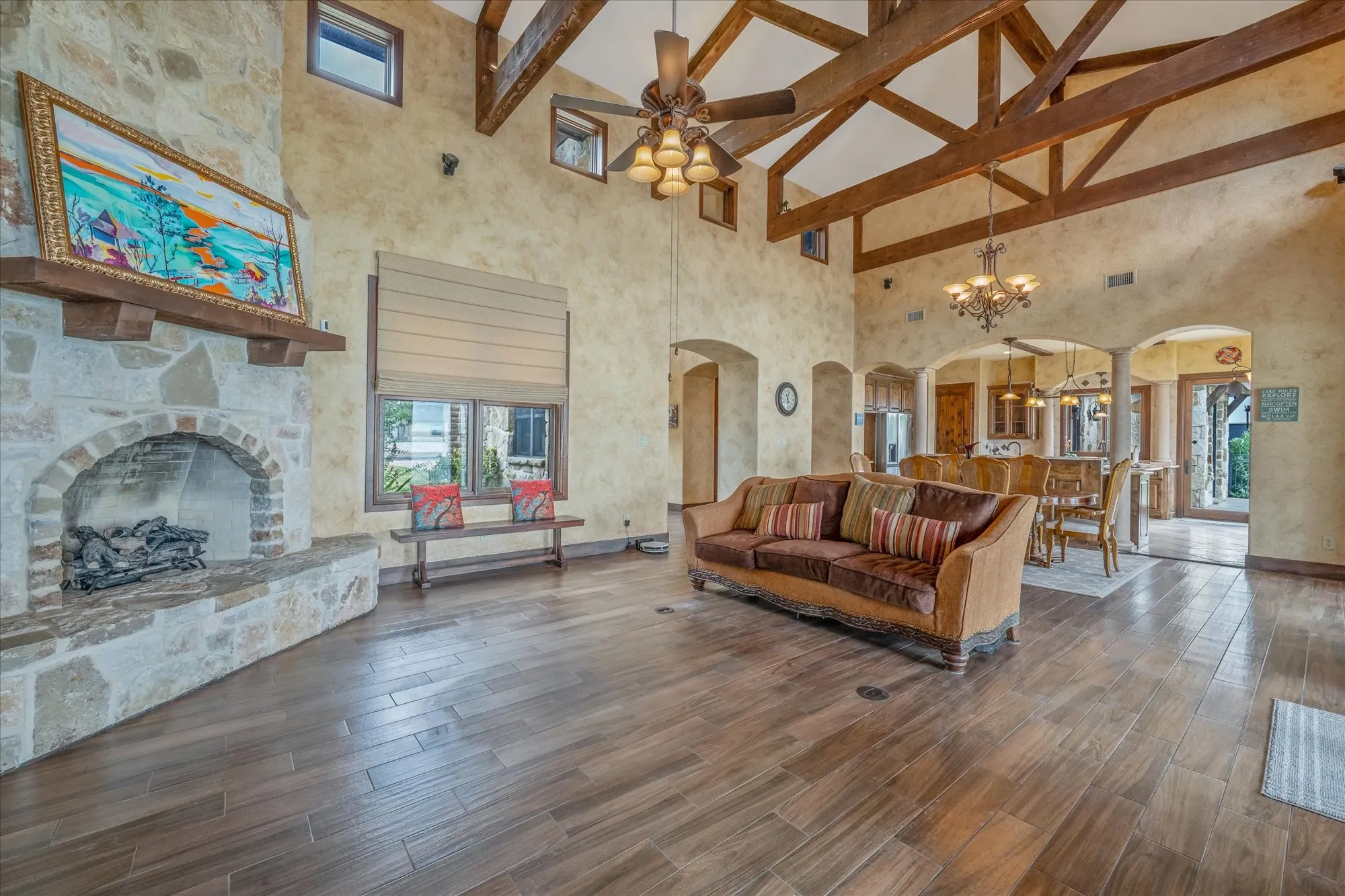 Vaulted ceiling with exposed wood beams, stone fireplace, wood-finish flooring, arched doorways, and a multi-light chandelier