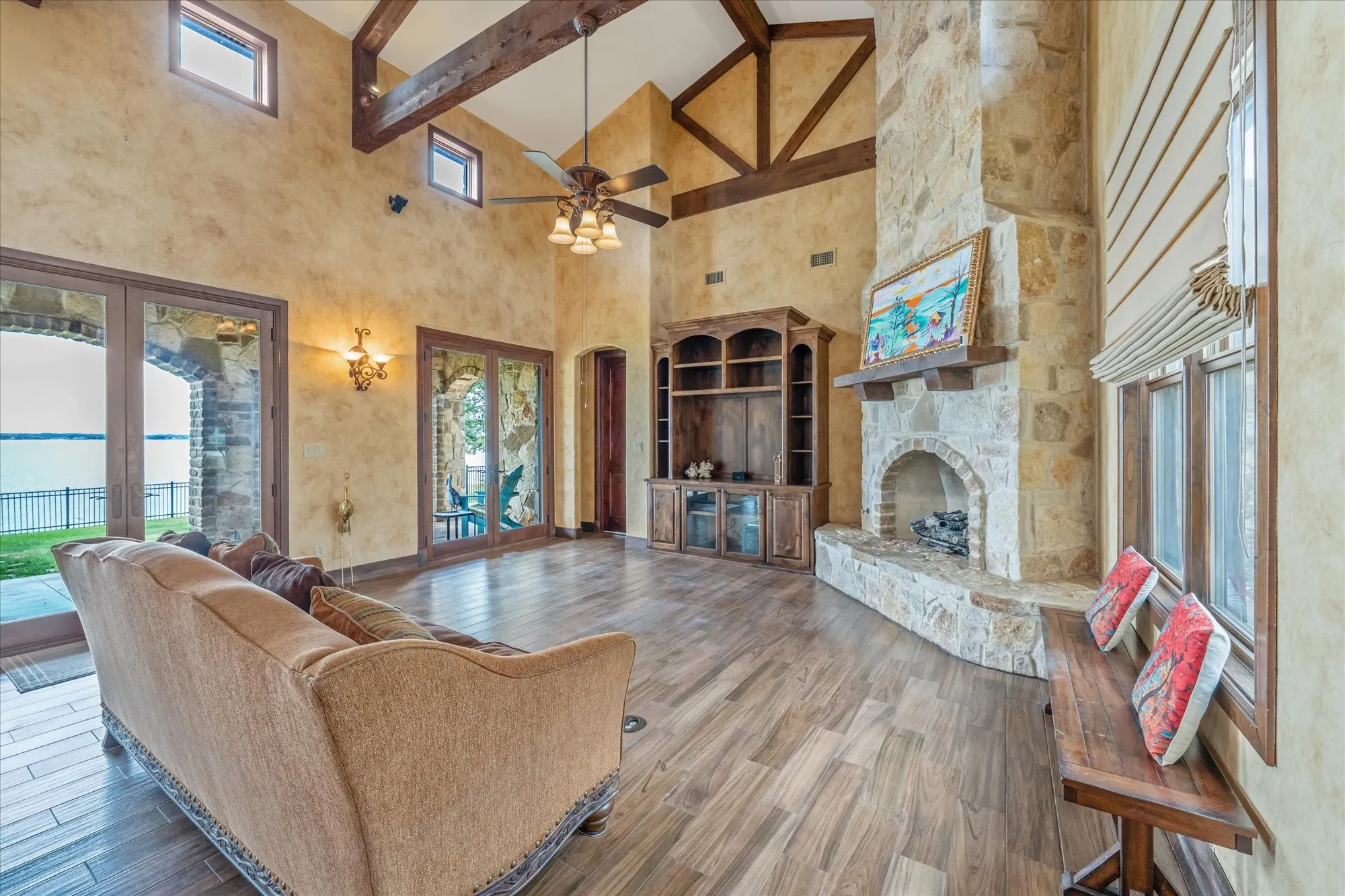 Vaulted ceiling with exposed wood beams, stone fireplace, wood-finish flooring, built-in entertainment center, and multiple glass French doors