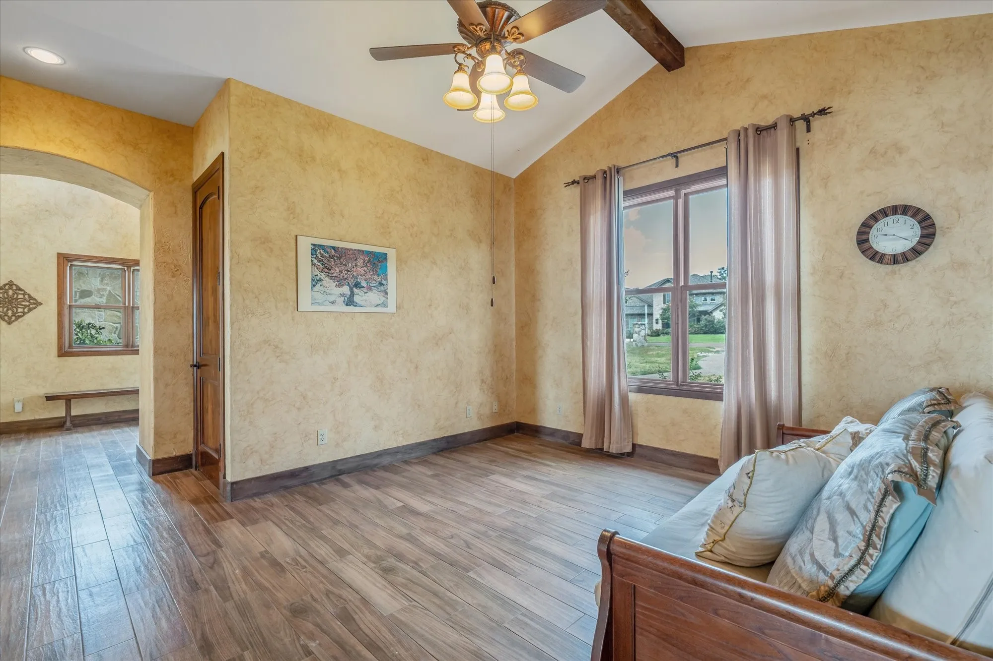 Vaulted ceiling with exposed wood beams, ceiling fan with integrated lighting, wood-finish flooring, and arched doorway