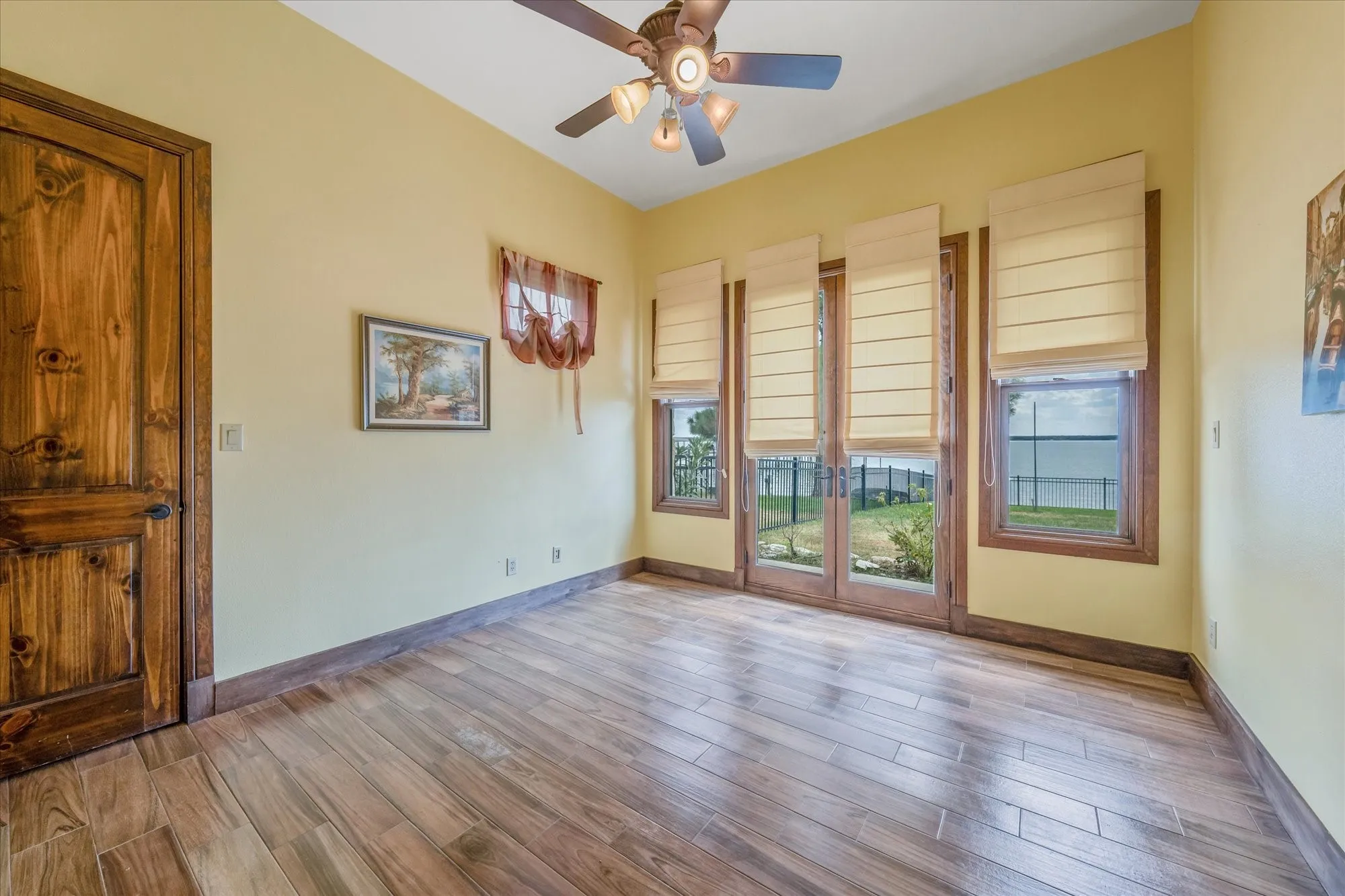 Wood-finish flooring, warm yellow walls, wood panel door, ceiling fan with light fixture, multiple windows with blinds