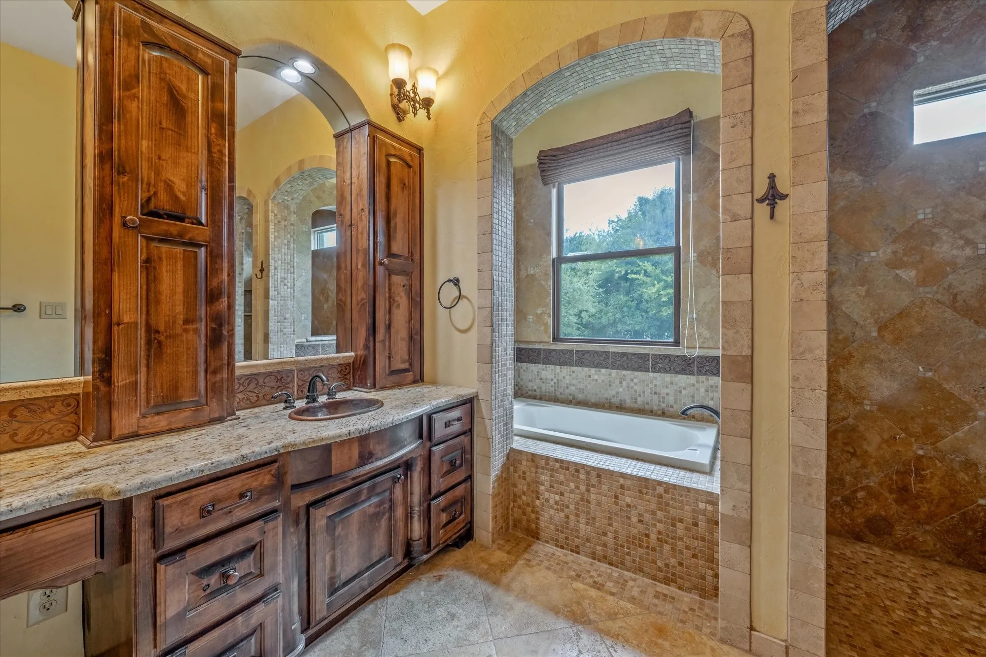 Granite-top vanity with integrated sink, rich wood-tone cabinetry, arched mirrors, and a built-in tub with mosaic tile surround