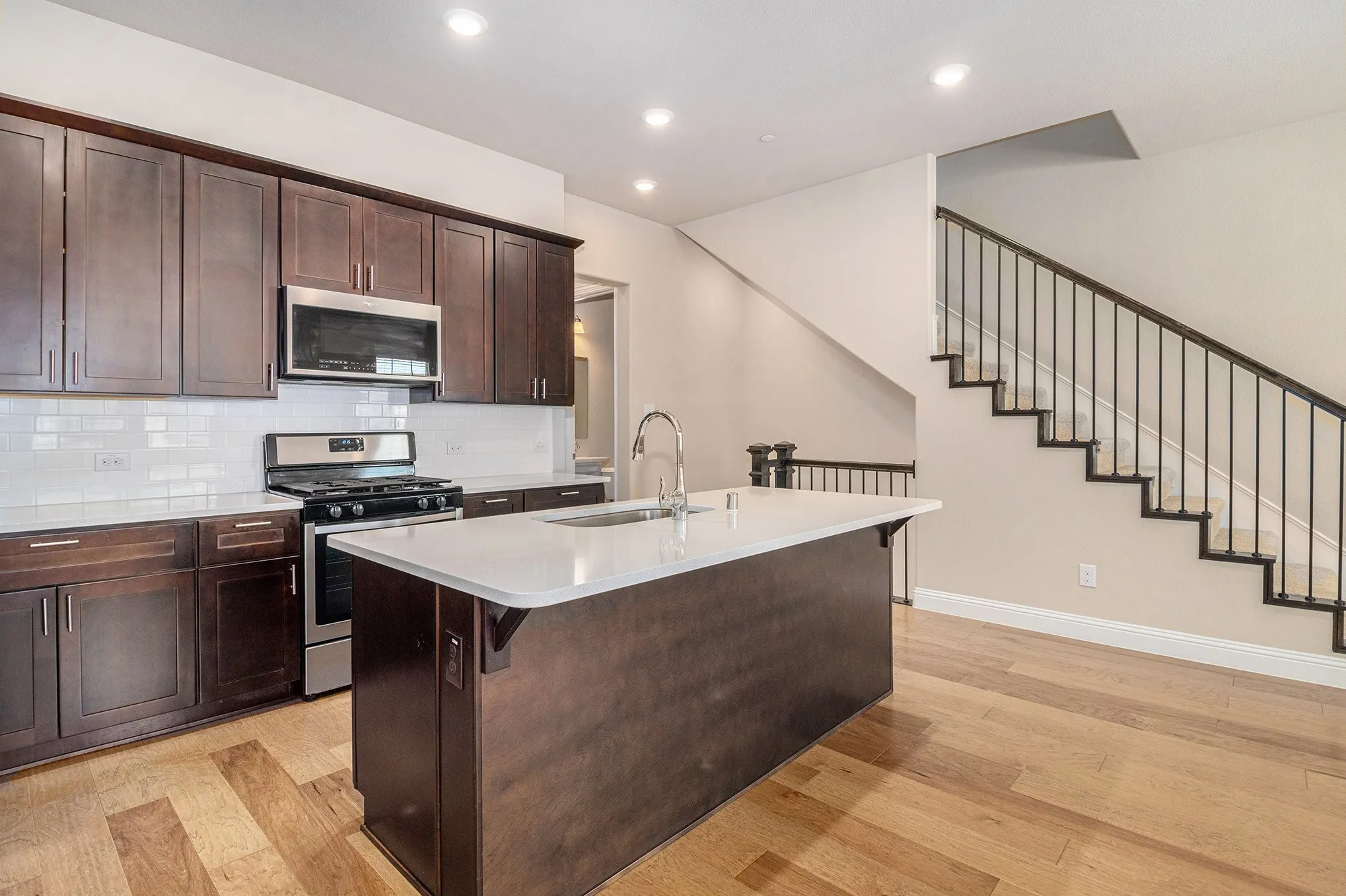 Kitchen island with an undermount sink, stainless steel appliances, dark wood cabinetry, white subway tile backsplash, and light wood flooring