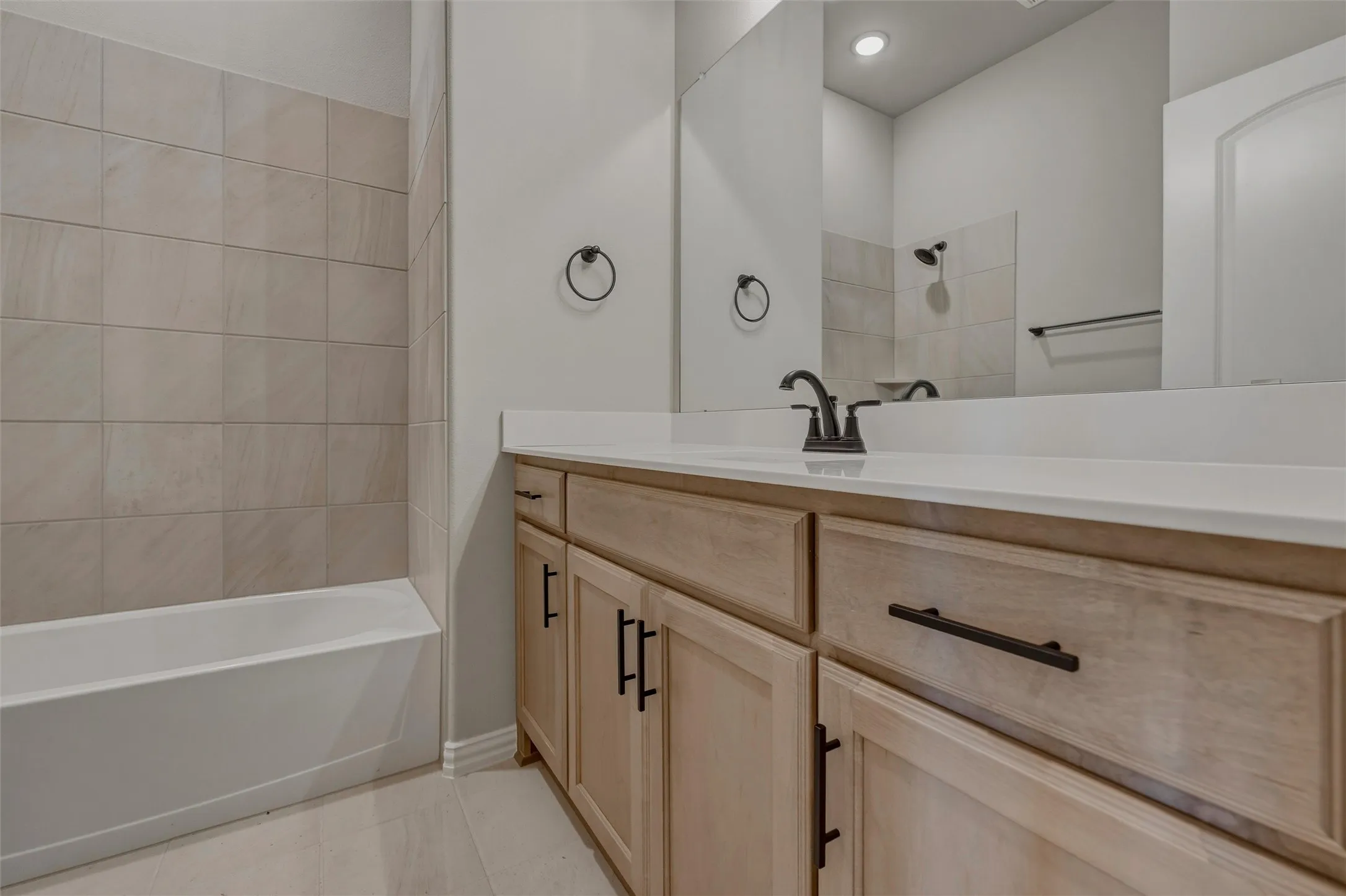 Bathroom featuring a light wood-finish vanity with dark hardware, a white countertop, and a built-in tub with neutral-toned tiled surround