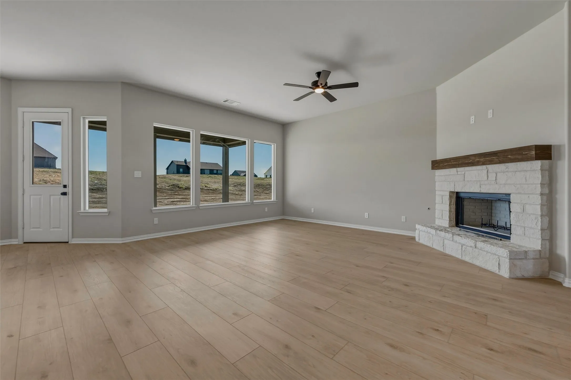 Light wood-finish flooring throughout, a white stone fireplace with a wood mantel, multiple large windows, and a ceiling fan