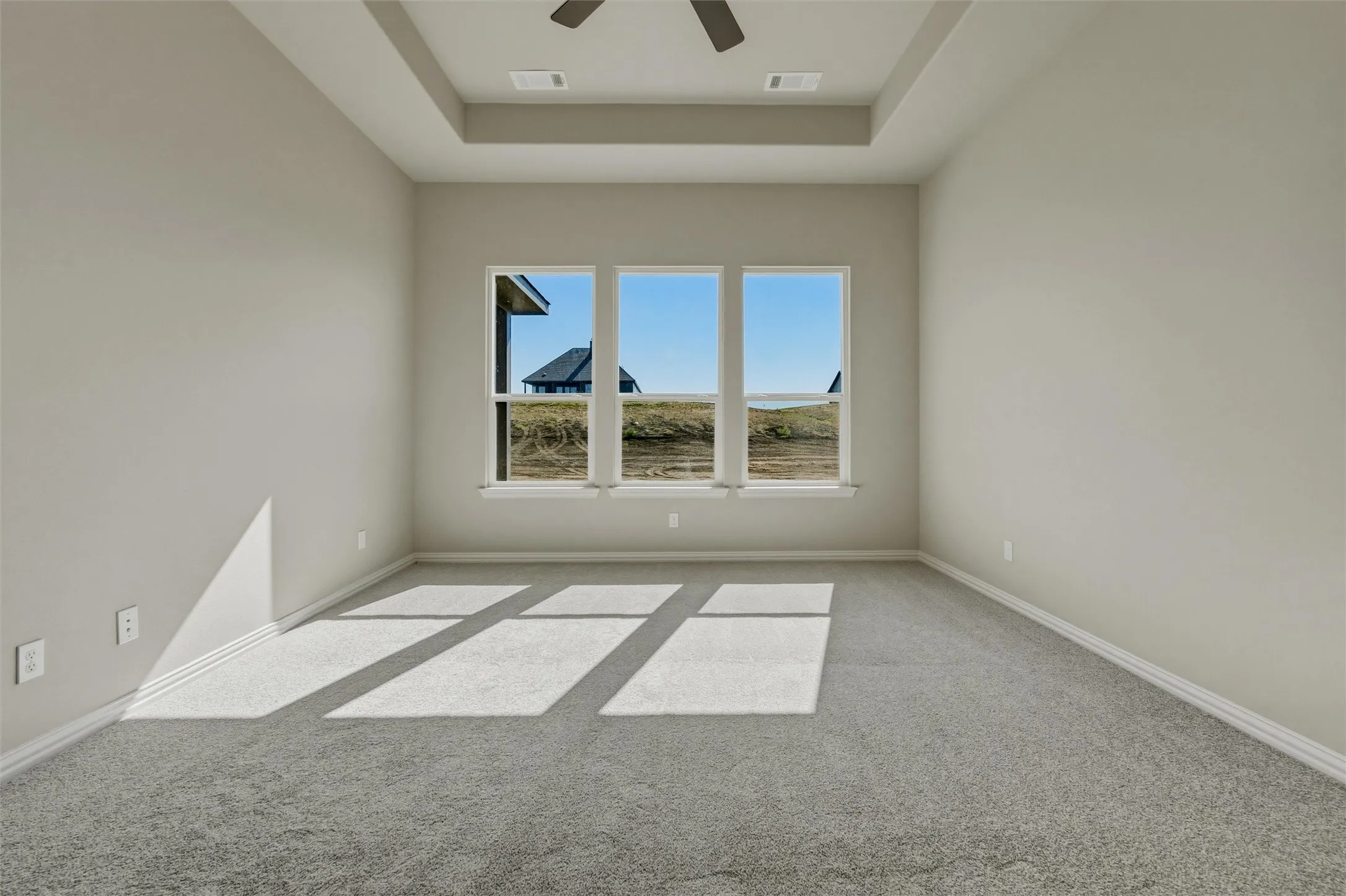 Tray ceiling with a ceiling fan, light gray wall paint, three large windows, white window trim, and gray carpet flooring