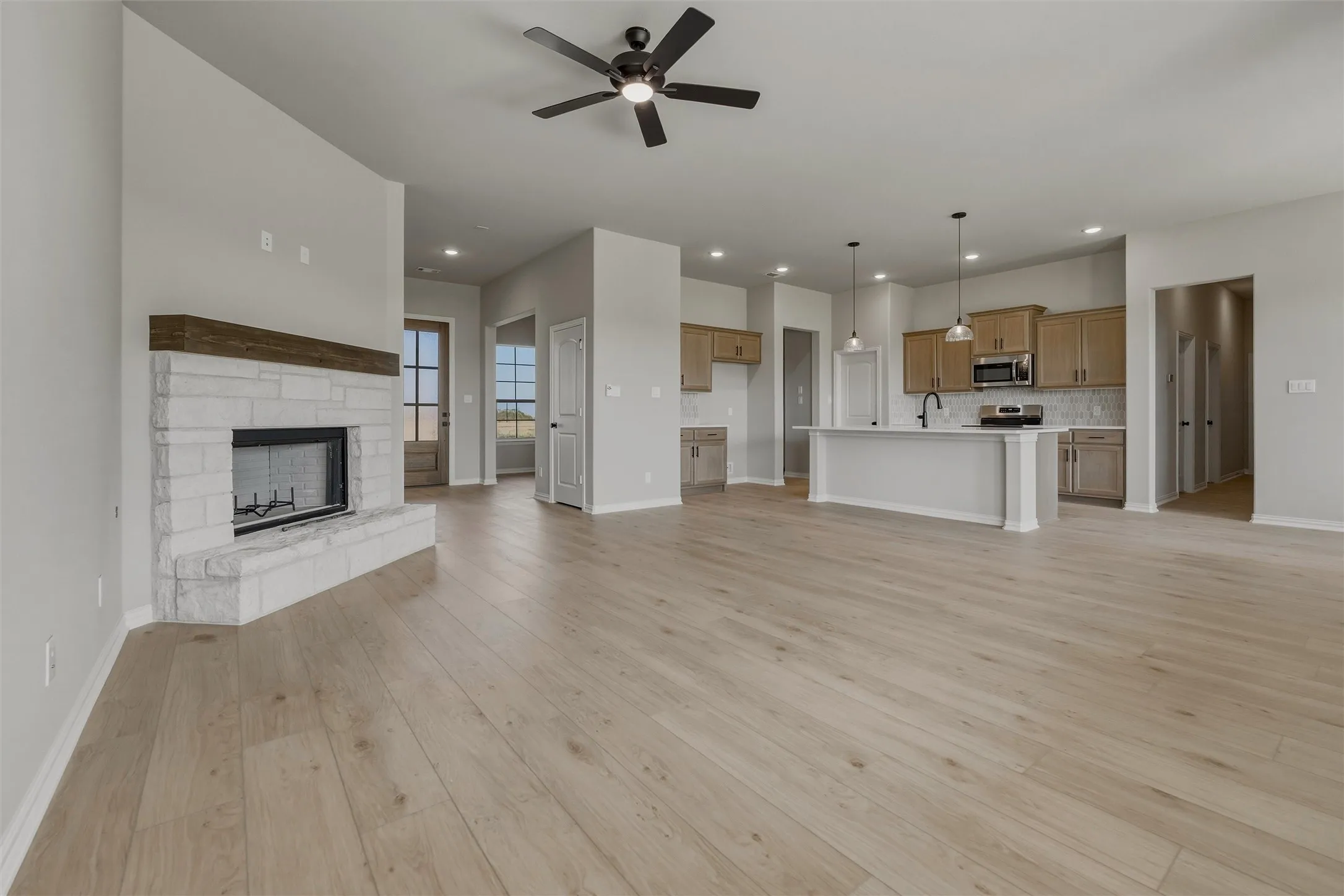 Spacious open-concept living area featuring light wood-finish flooring, a stone-clad fireplace with a wood mantel, and a ceiling fan
