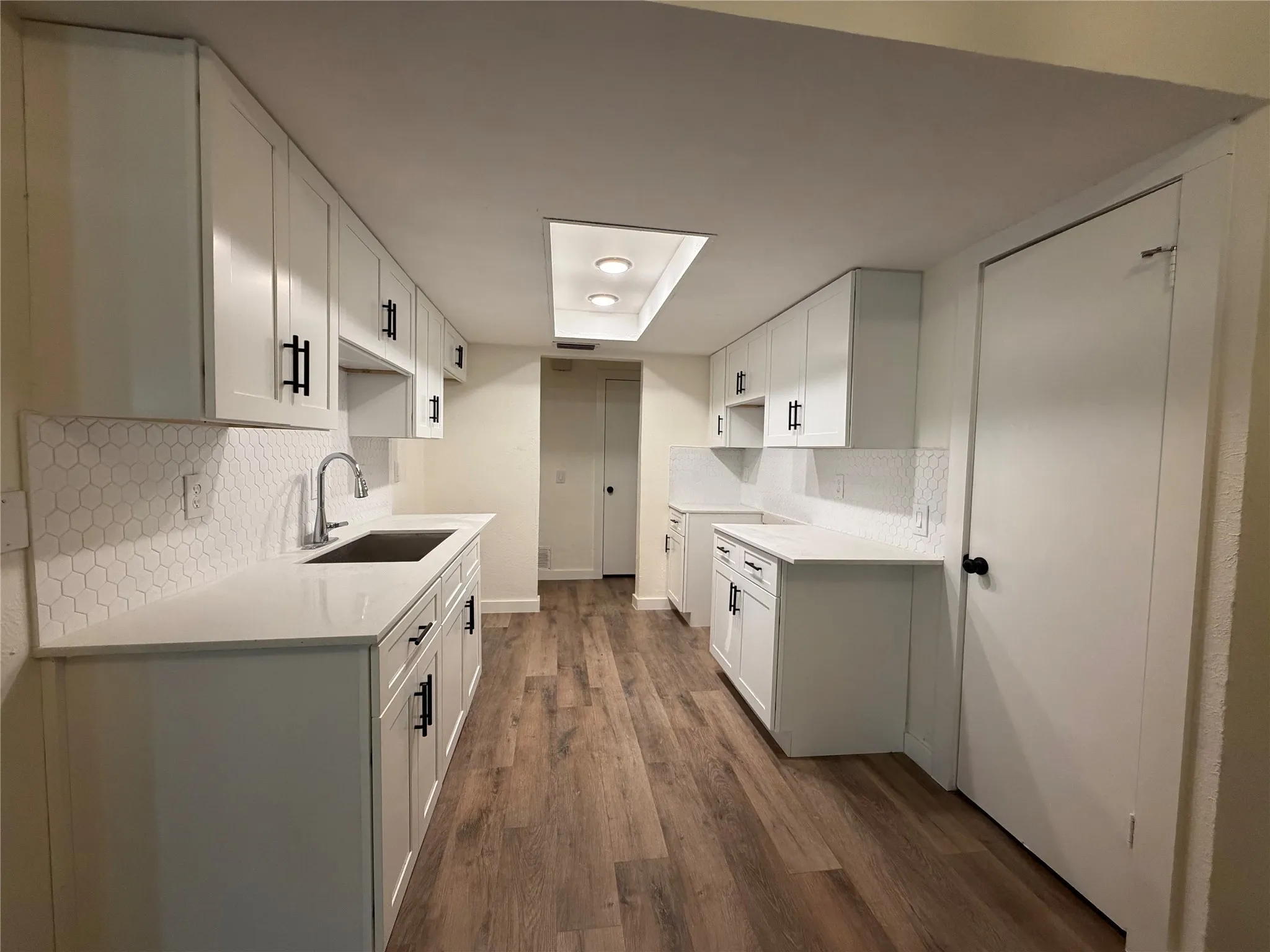 White shaker-style cabinetry with matte black hardware, hexagonal tile backsplash, stainless steel sink with gooseneck faucet, light-toned countertops, and wood-finish flooring