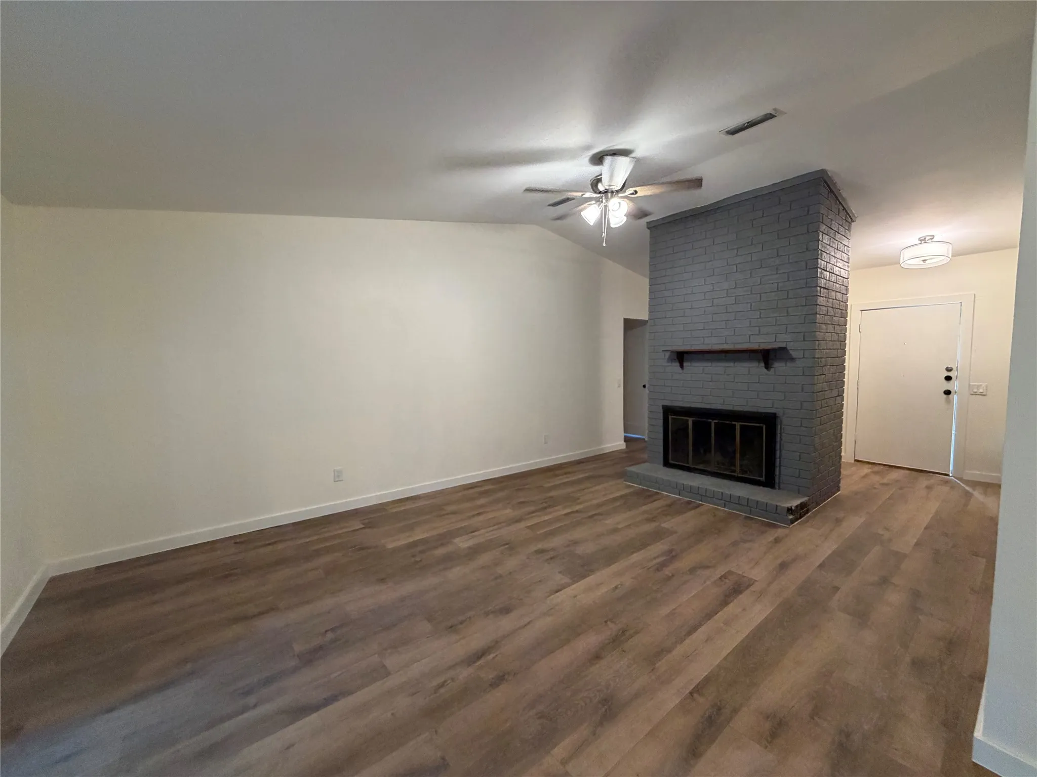 Wood-finish flooring throughout, vaulted ceiling, painted brick fireplace with a wood mantle, and a ceiling fan with integrated lighting