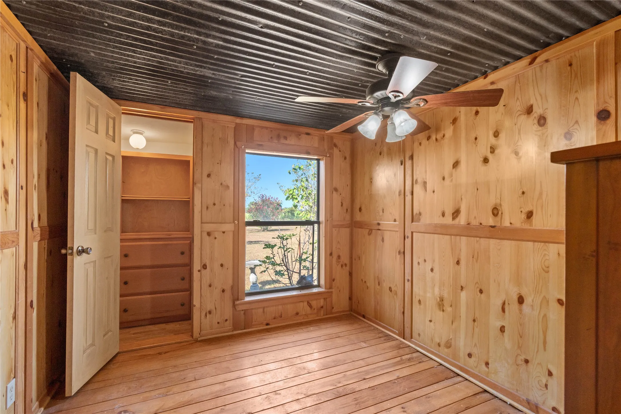 Wood-paneled walls, corrugated metal ceiling, wood-finish flooring, built-in closet storage, and a ceiling fan with integrated lighting