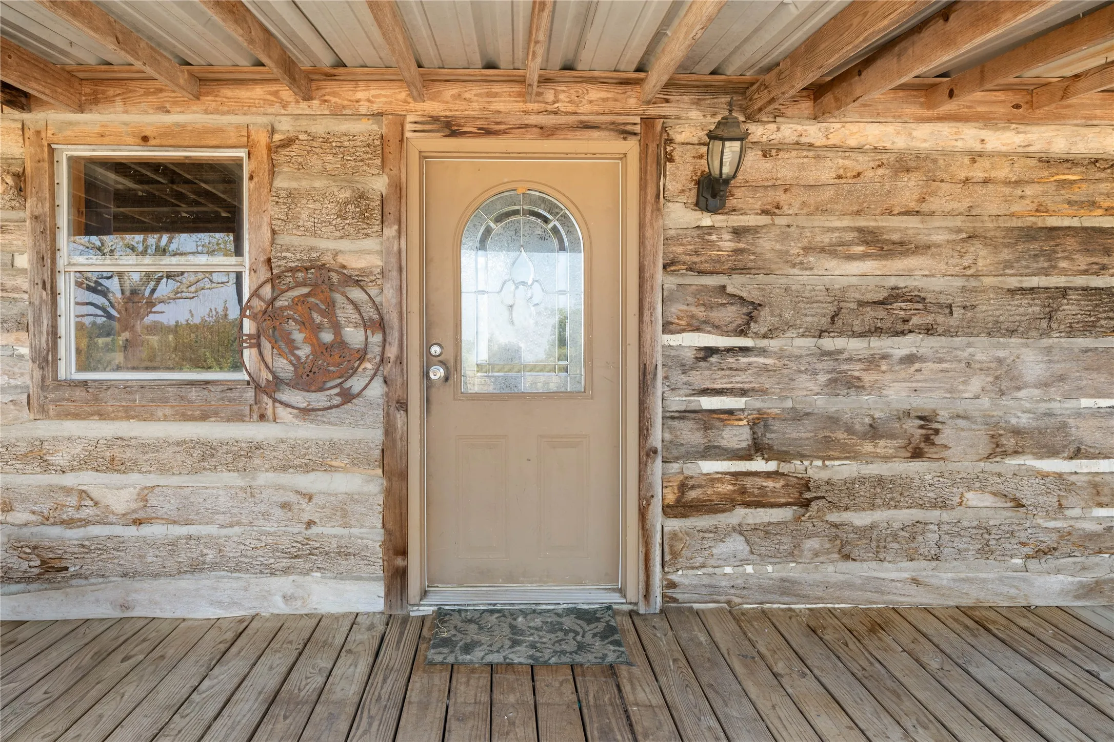 Wood-plank exterior wall with a wood-plank porch floor, featuring a front door with an arched glass insert, an exterior wall-mounted light fixture, and a single-pane window