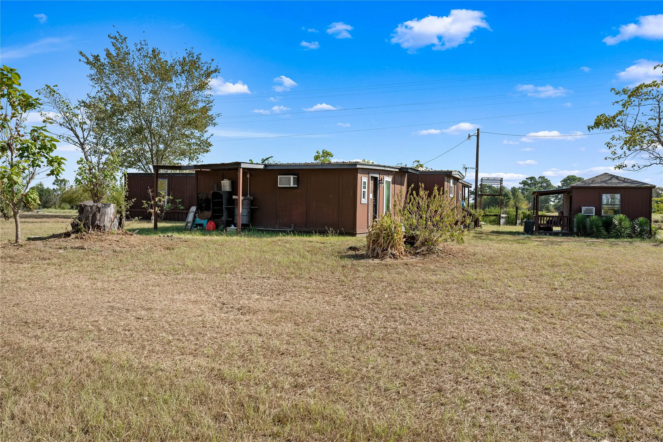 Brown-sided structures on a grassy lot, featuring a covered porch and mature trees