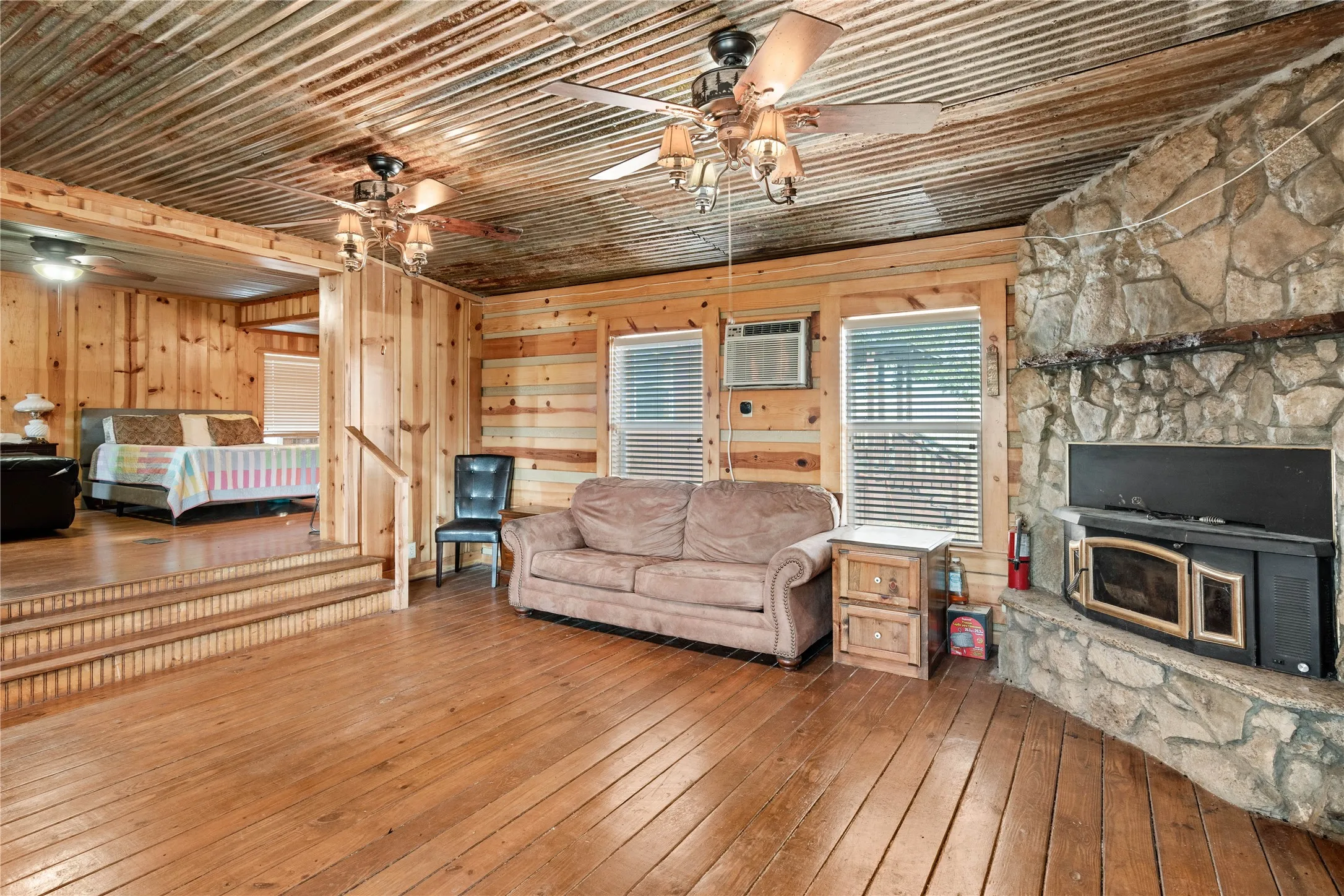 Wood-finish flooring throughout, corrugated metal ceiling, stone-clad fireplace with insert, and wood-paneled walls