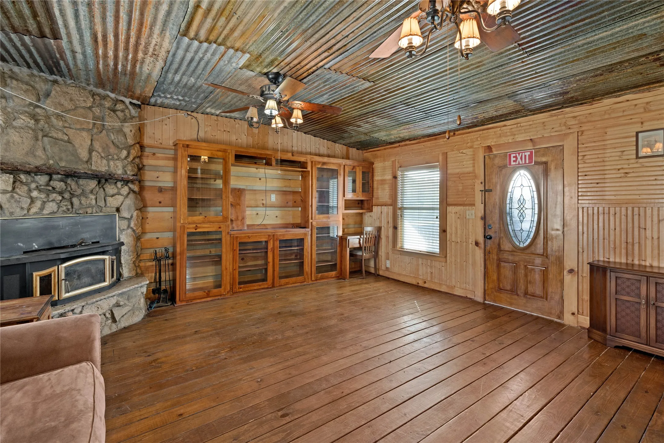 Corrugated metal ceiling, wood-finish flooring, stone fireplace with a wood-burning insert, built-in cabinetry, and a wood entry door with an oval glass insert