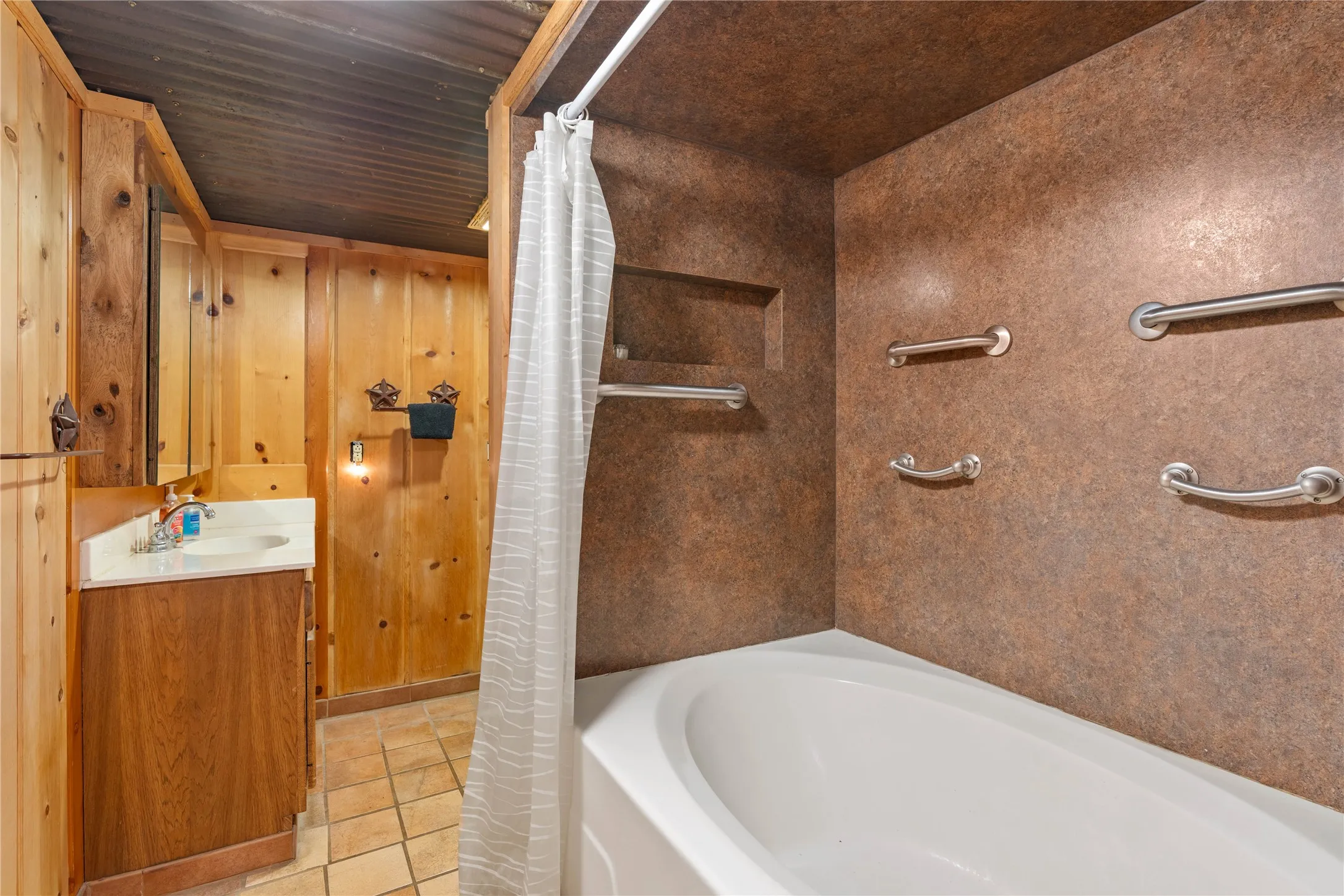 Bathtub with integrated wall-mounted grab bars and a built-in shower niche, complemented by wood paneling, a wood-finish vanity, and a corrugated metal ceiling