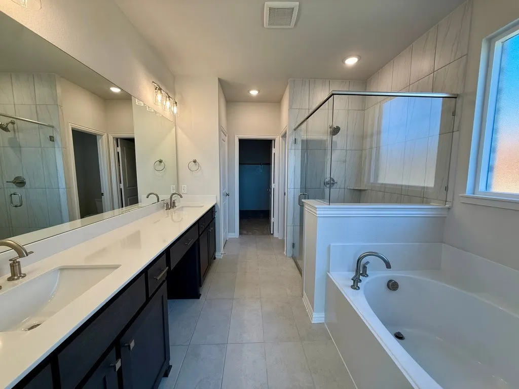 Dual vanity with integrated sinks and dark cabinetry, large soaking tub, walk-in shower with glass enclosure, tiled shower walls, and recessed lighting