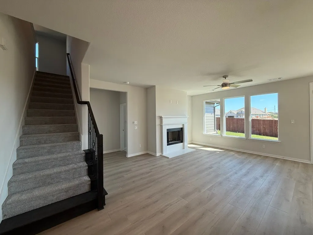 Wood-finish flooring, white mantel fireplace, three large windows, ceiling fan, and carpeted stairs with a dark wood and iron railing