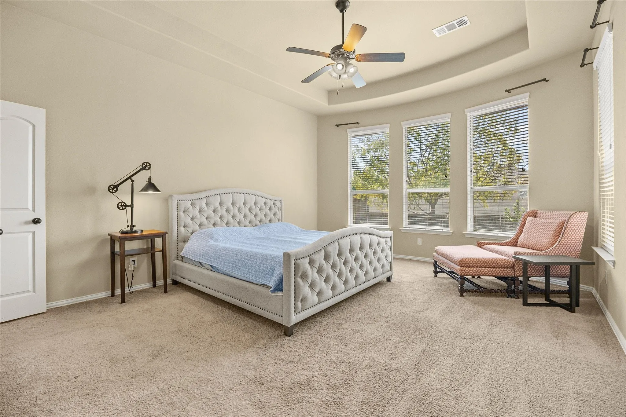 Tray ceiling with a ceiling fan, three large windows with blinds, and neutral carpeting