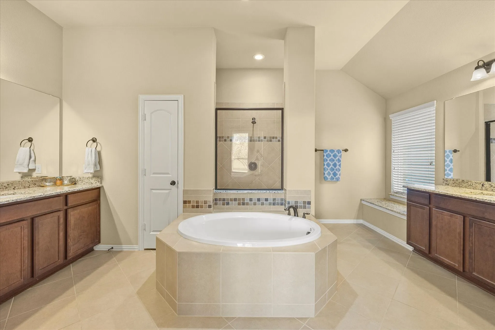 Tile flooring, built-in soaking tub, separate glass-enclosed shower, dual vanities with granite countertops, and a window with a built-in bench