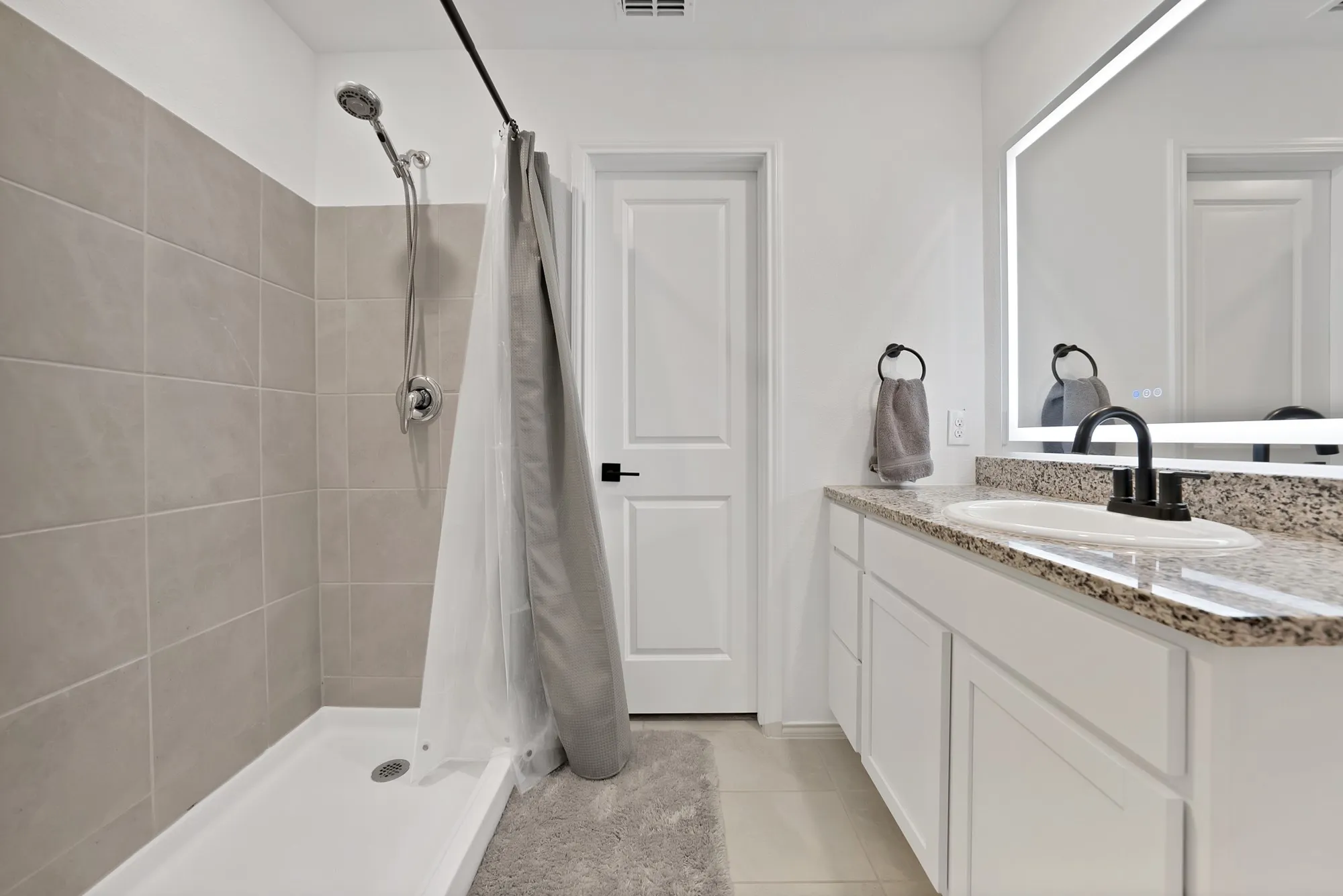 Bathroom featuring a shower with light-toned tile surround, white vanity with stone-finish countertop, undermount sink, matte black faucet, and an illuminated mirror