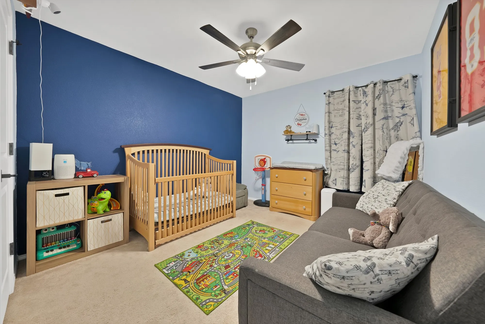 Versatile dual-space room featuring a wood-slat crib with natural finish, coordinating wood dresser with multiple drawers, built-in shelving with cubbies for storage, ceiling fan with integrated lighting, and neutral carpet flooring