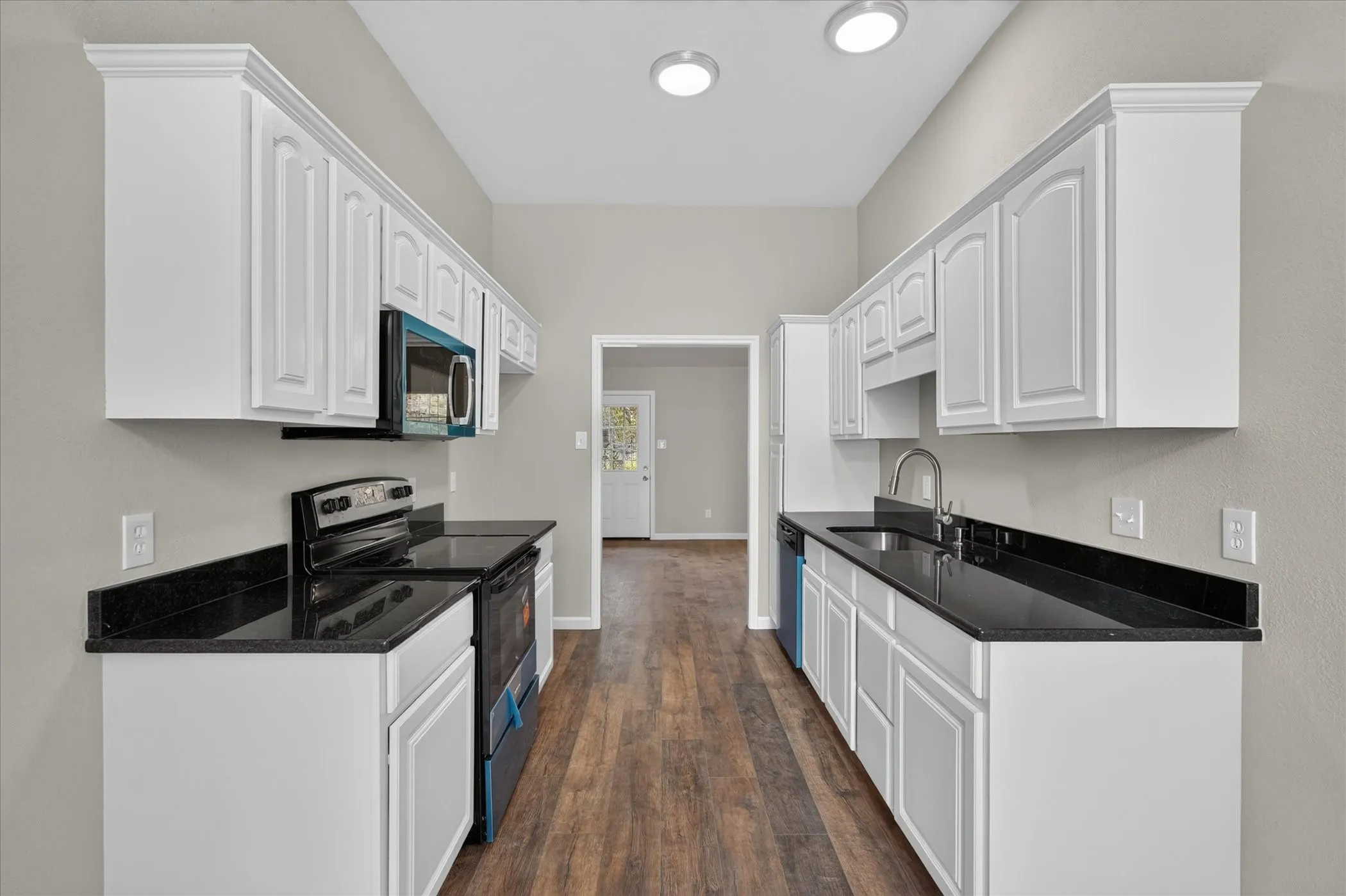 White cabinetry with crown molding, black countertops, stainless steel sink with gooseneck faucet, microwave, and wood-finish flooring