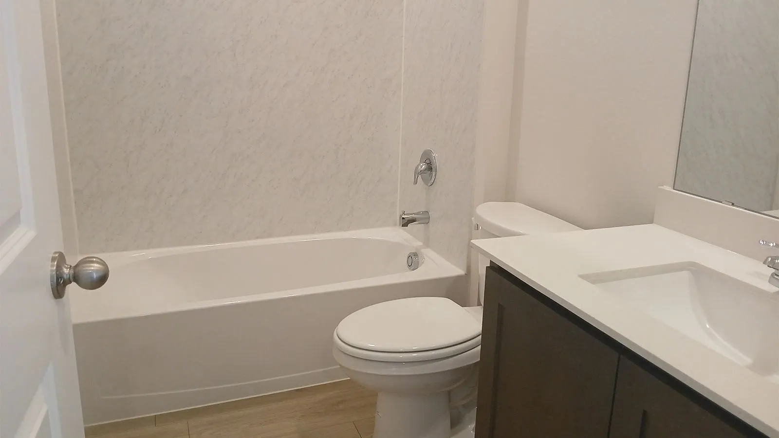 White bathtub and shower surround with chrome fixtures, wood-finish flooring, dark wood-finish vanity with white countertop, and integrated sink