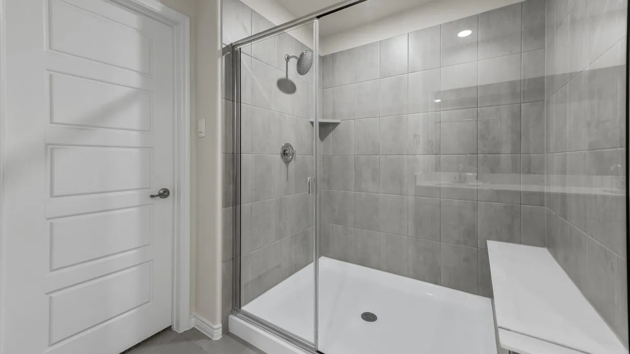 Glass-enclosed shower with a rainfall showerhead, handheld sprayer, built-in shelving, integrated bench, and gray tile surround